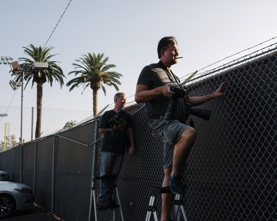 Two men stand on a ladder against a chain-link fence with palm trees visible in the background during daytime.