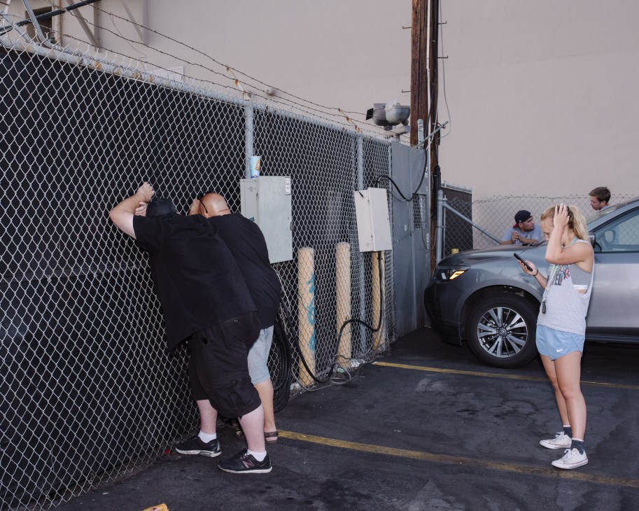 Paparazzi photographer shooting over a chain-link fence while a woman holds up a phone nearby.
