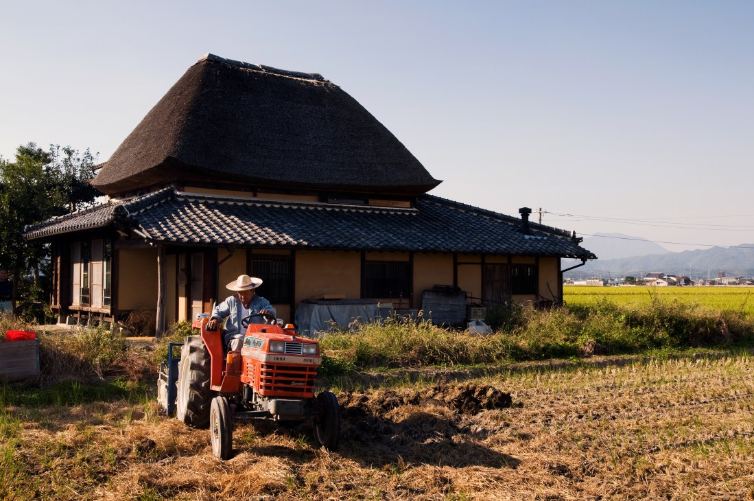 A farmer operates a red tractor in front of a traditional Japanese farmhouse with thatched roof in a rural rice field landscape.