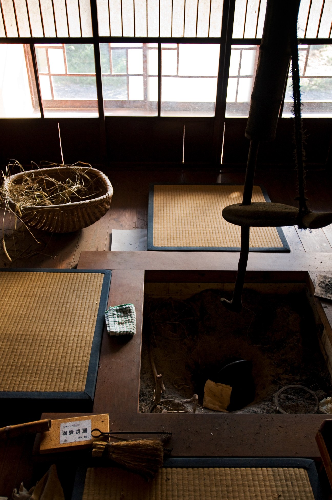 Interior of a traditional Japanese minka farmhouse with tatami mat flooring, wooden beams, paper screens, and an open hearth with basket and cooking implements.