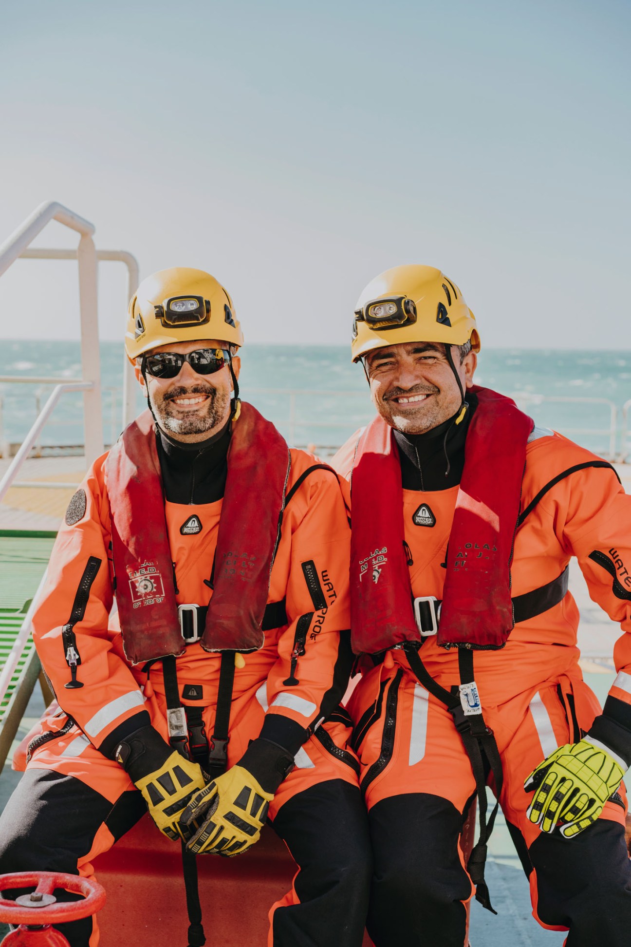 Two rescue divers in yellow helmets and orange survival suits smile at the camera on the deck of a ship with the ocean behind them.