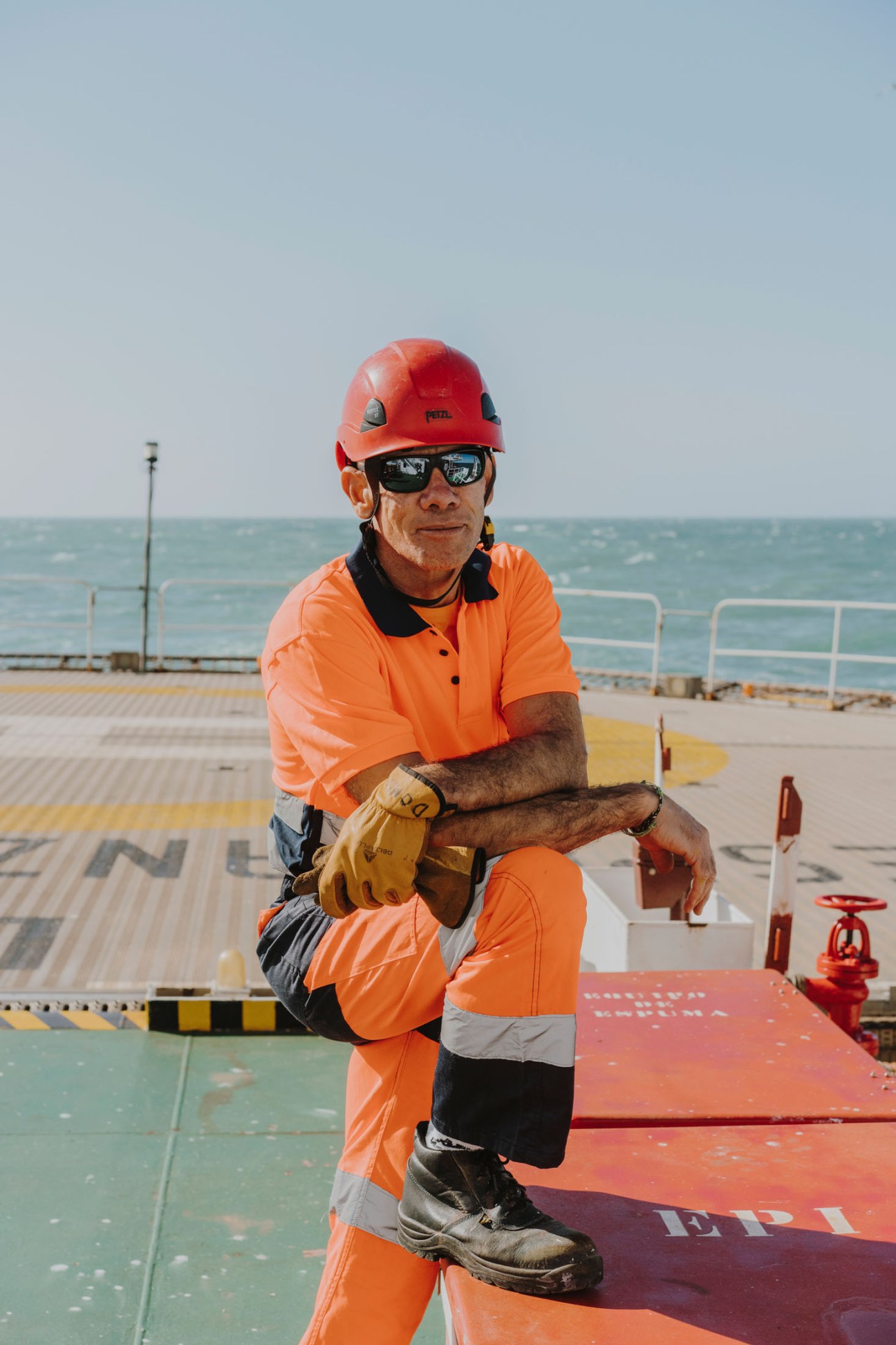 A crew member in orange safety gear and red hard hat squats on the ship's deck during a rescue drill.