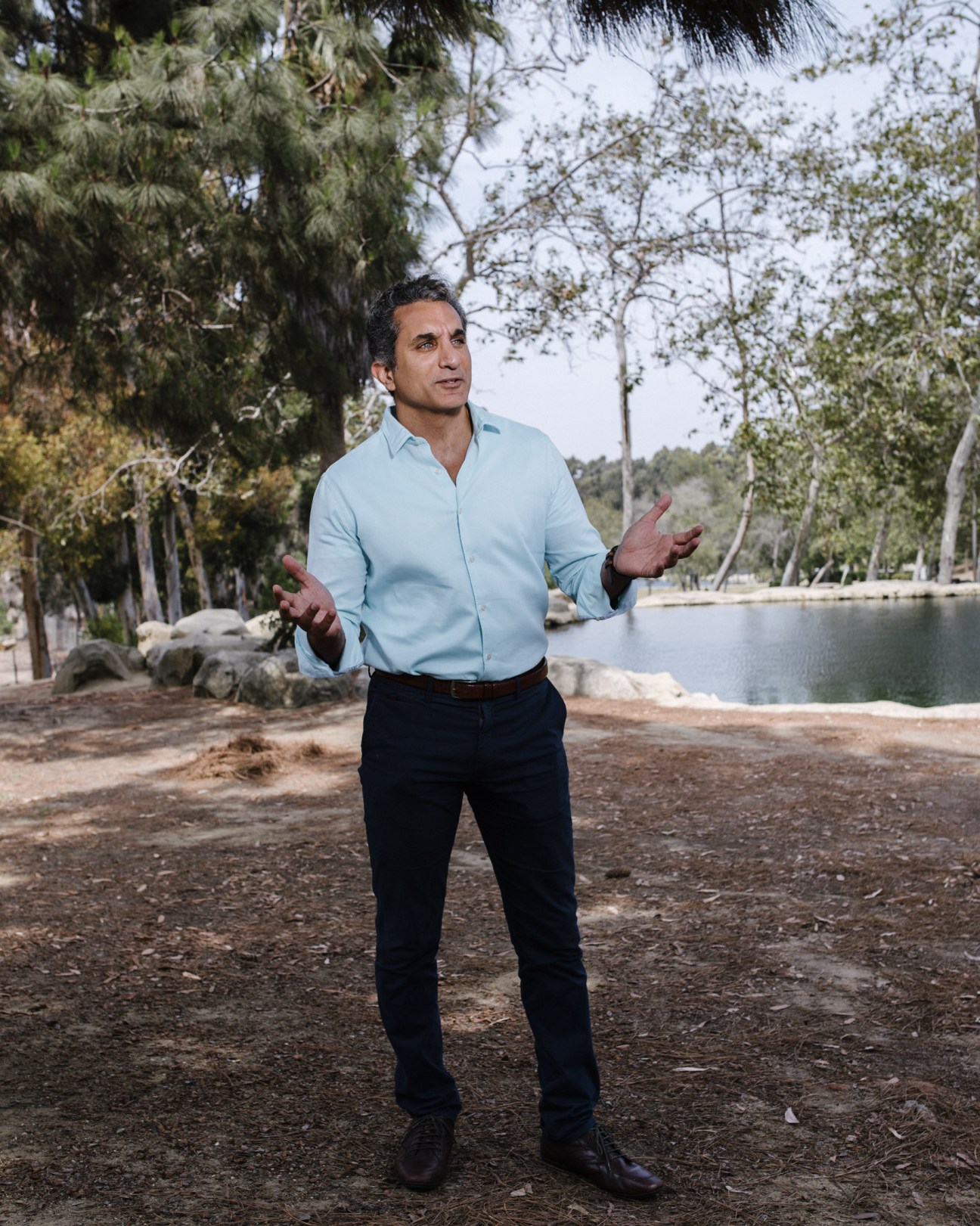 Bassem Youssef standing outdoors by a lake surrounded by trees, wearing a light blue shirt and dark pants.