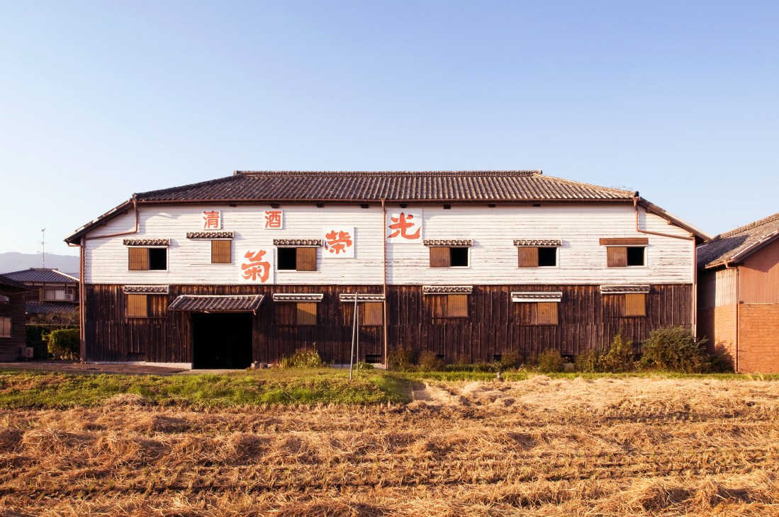 Traditional Japanese sake brewery with white upper facade, dark wooden lower level, and tiled roof with Japanese characters.