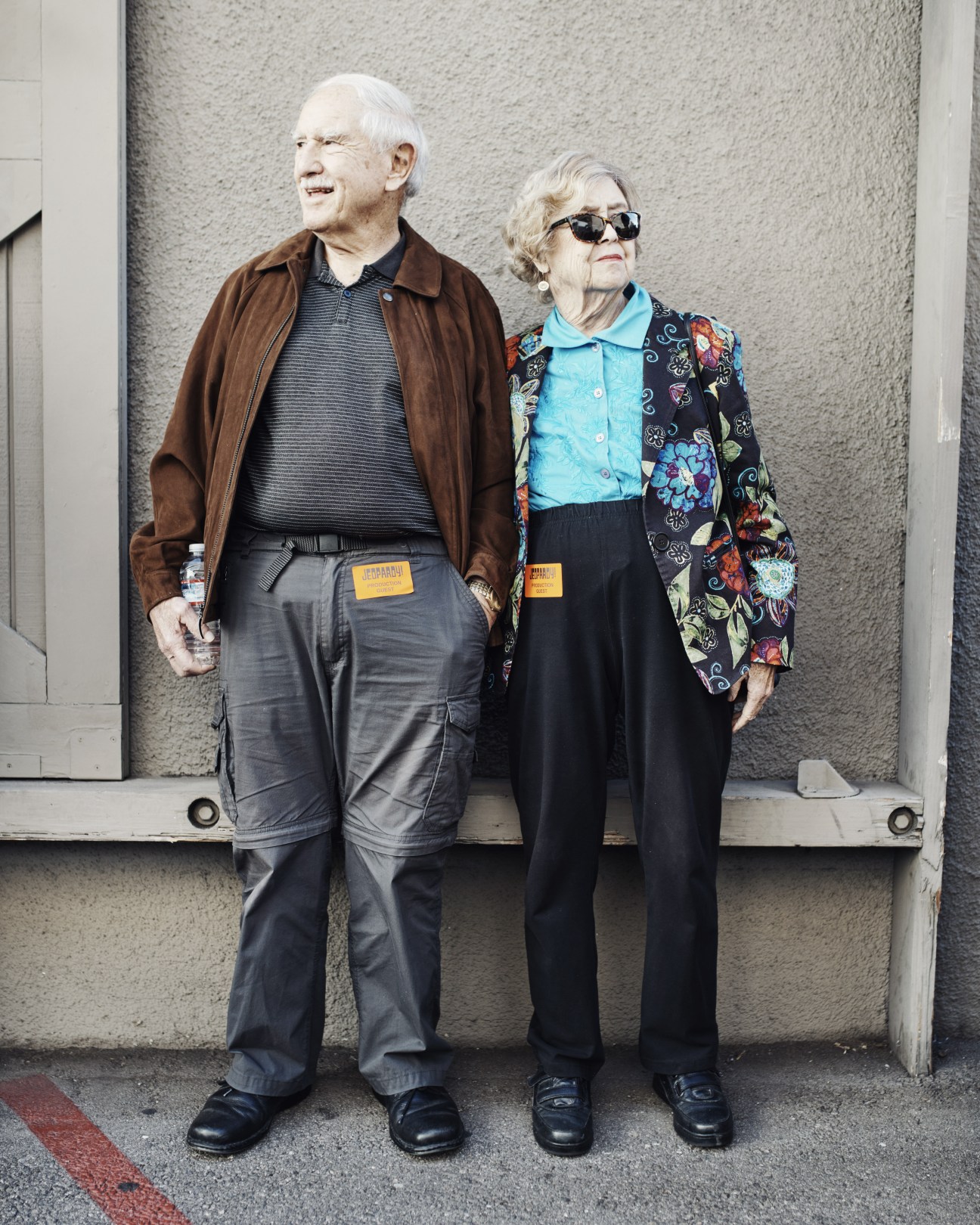Two elderly people wearing contestant badges stand outside a studio building.