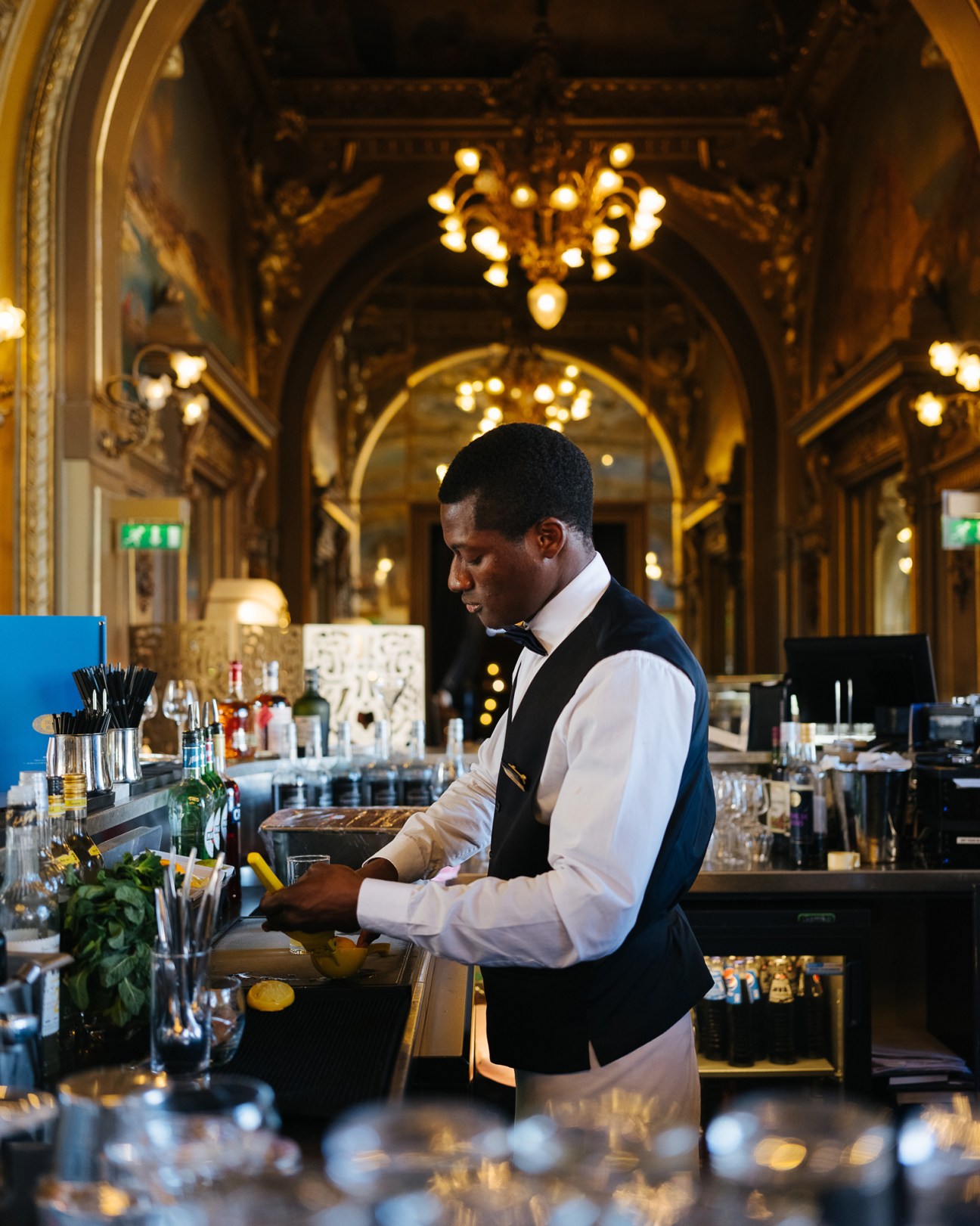 Bartender in formal attire mixing drinks at Le Train Bleu's ornate bar with gilded architecture and chandeliers.