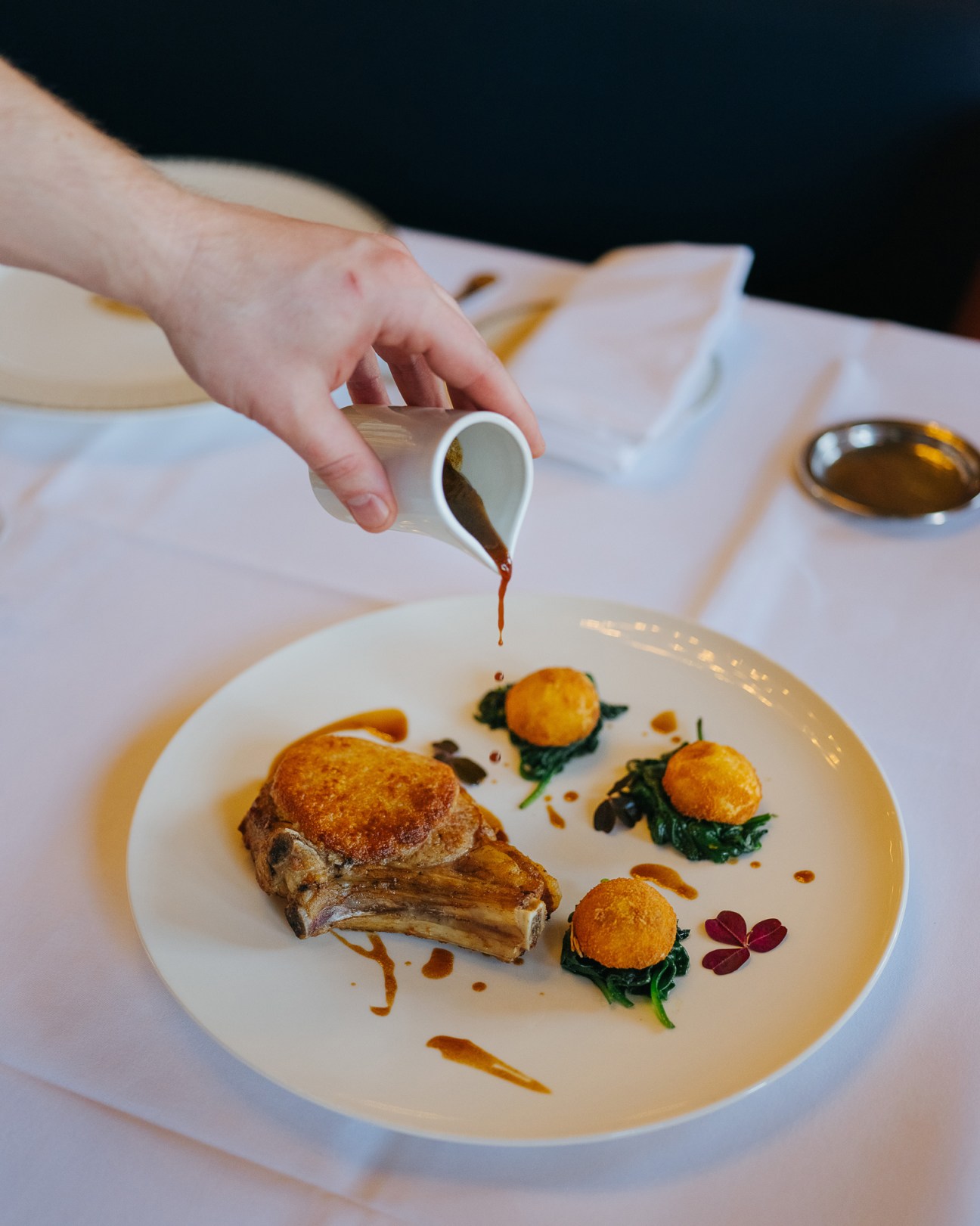 A hand drizzles sauce over a plated veal chop with golden croquettes and microgreens at Le Train Bleu restaurant.