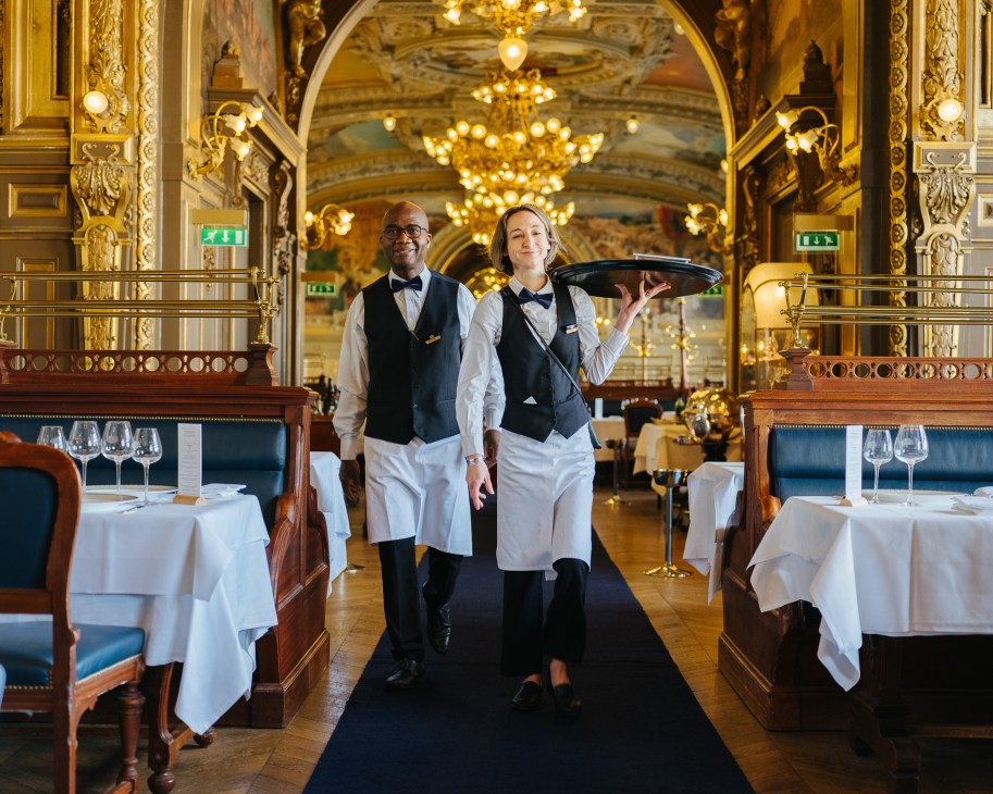 Two waiters in formal attire walking through Le Train Bleu's ornate dining room with gilded walls and chandeliers.