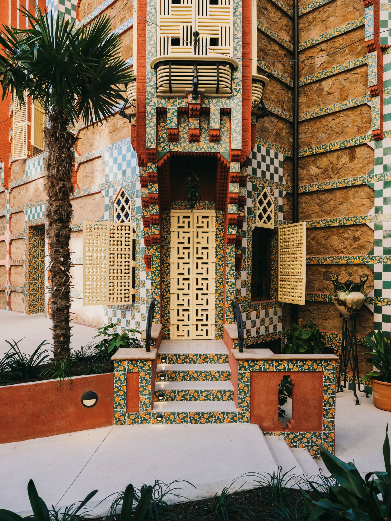Casa Vicens exterior entrance with colorful hand-painted ceramic tiles, geometric patterns, and palm plants framing the ornate doorway.