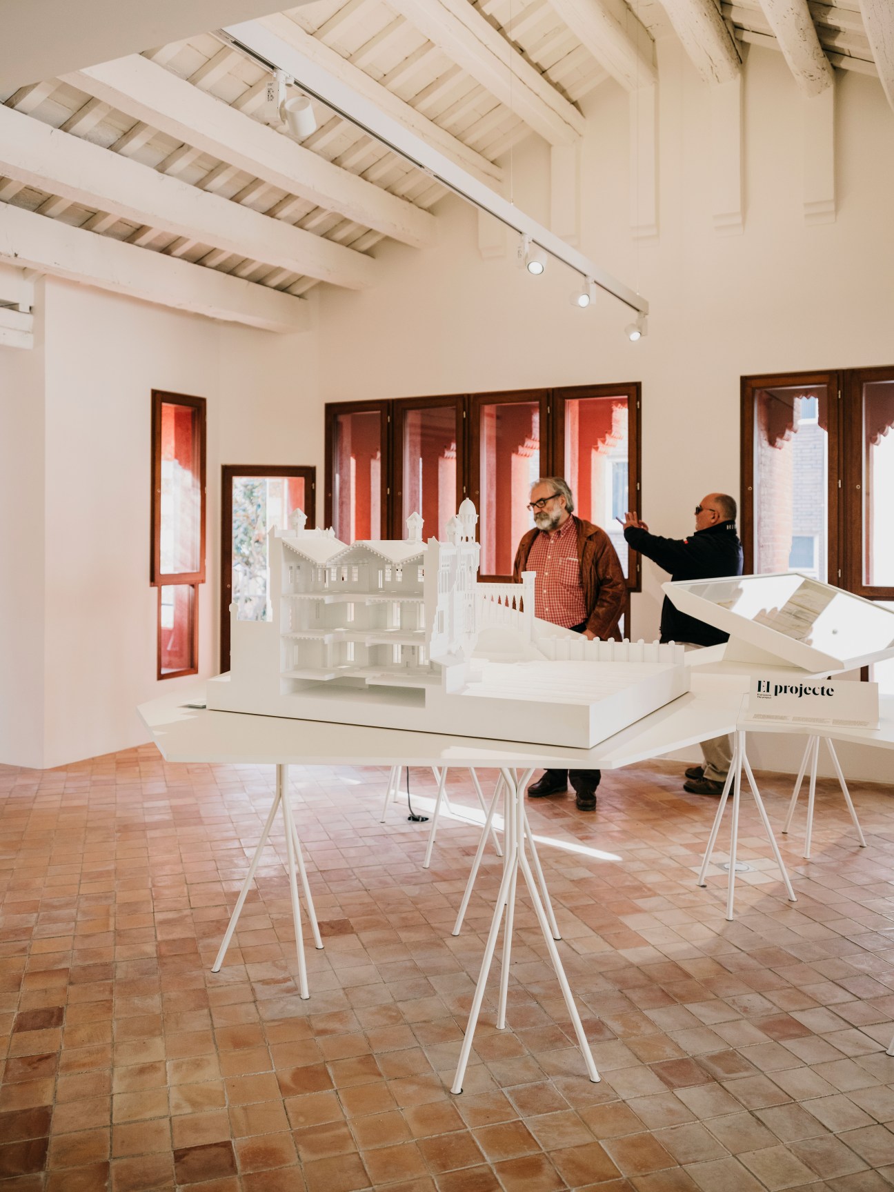 Two visitors examine architectural models in a modern white gallery space with terracotta flooring and exposed wooden ceiling beams.