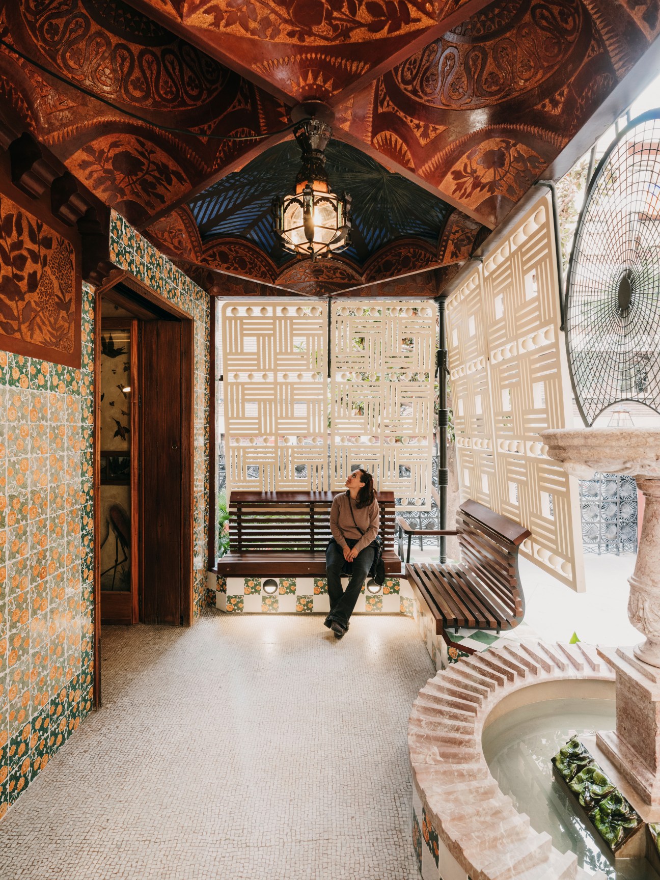 Woman sitting on bench in Casa Vicens terrace with ornate orange ceiling, colorful tiles and geometric lattice screens.
