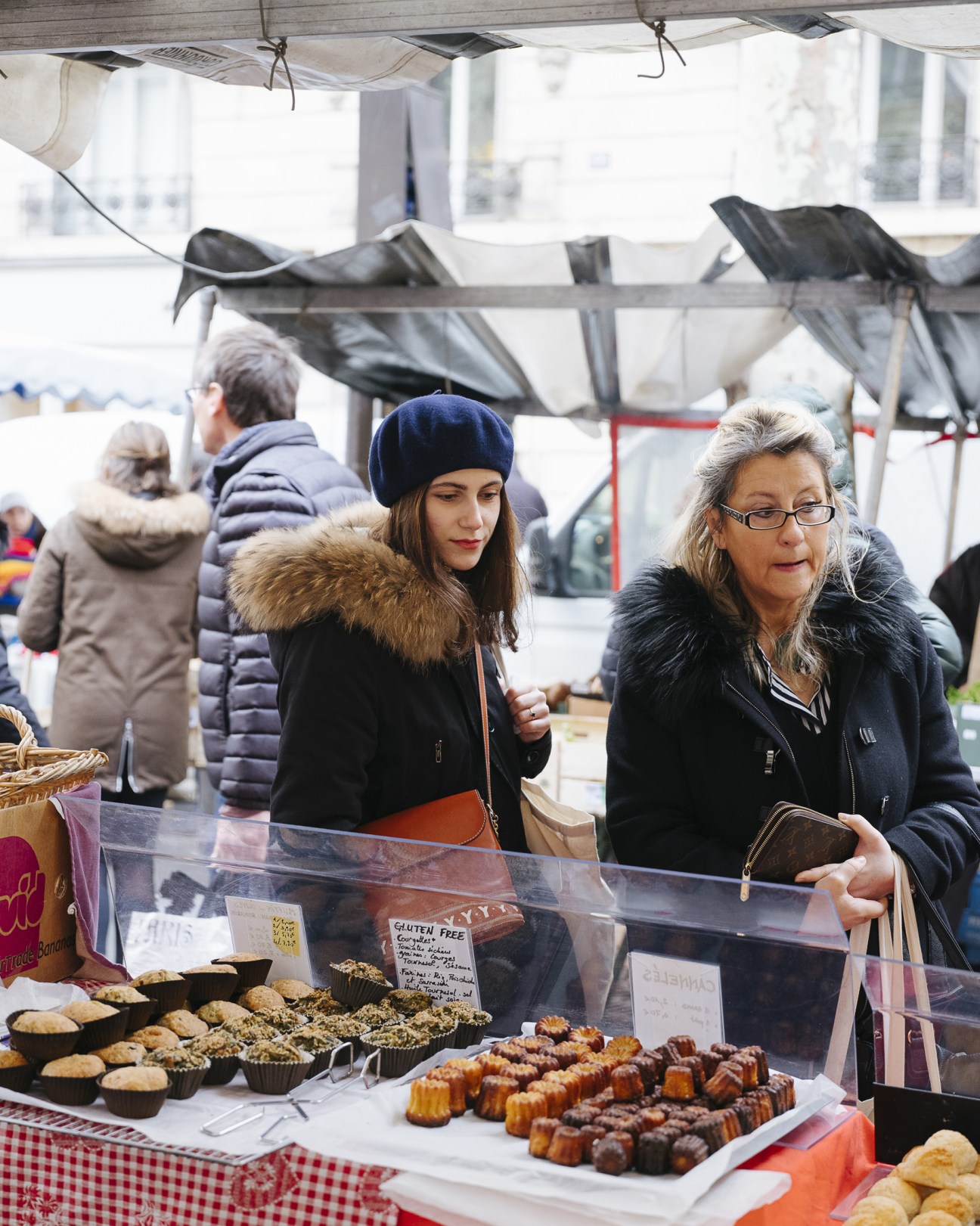 Two women browse baked goods and pastries at a market stall under a white tent.