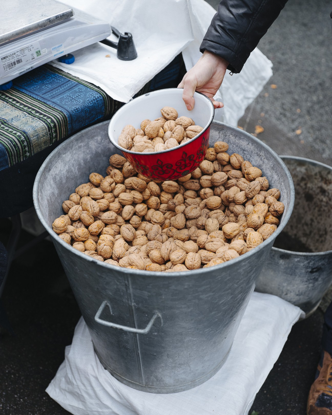 Hand scooping walnuts from a red and white bowl into a large metal bucket filled with nuts at a market stall.