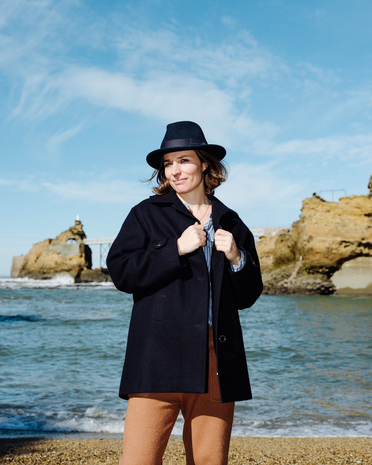 Marie Guyot stands on a beach wearing a black coat and hat, with rocky cliffs visible behind her.