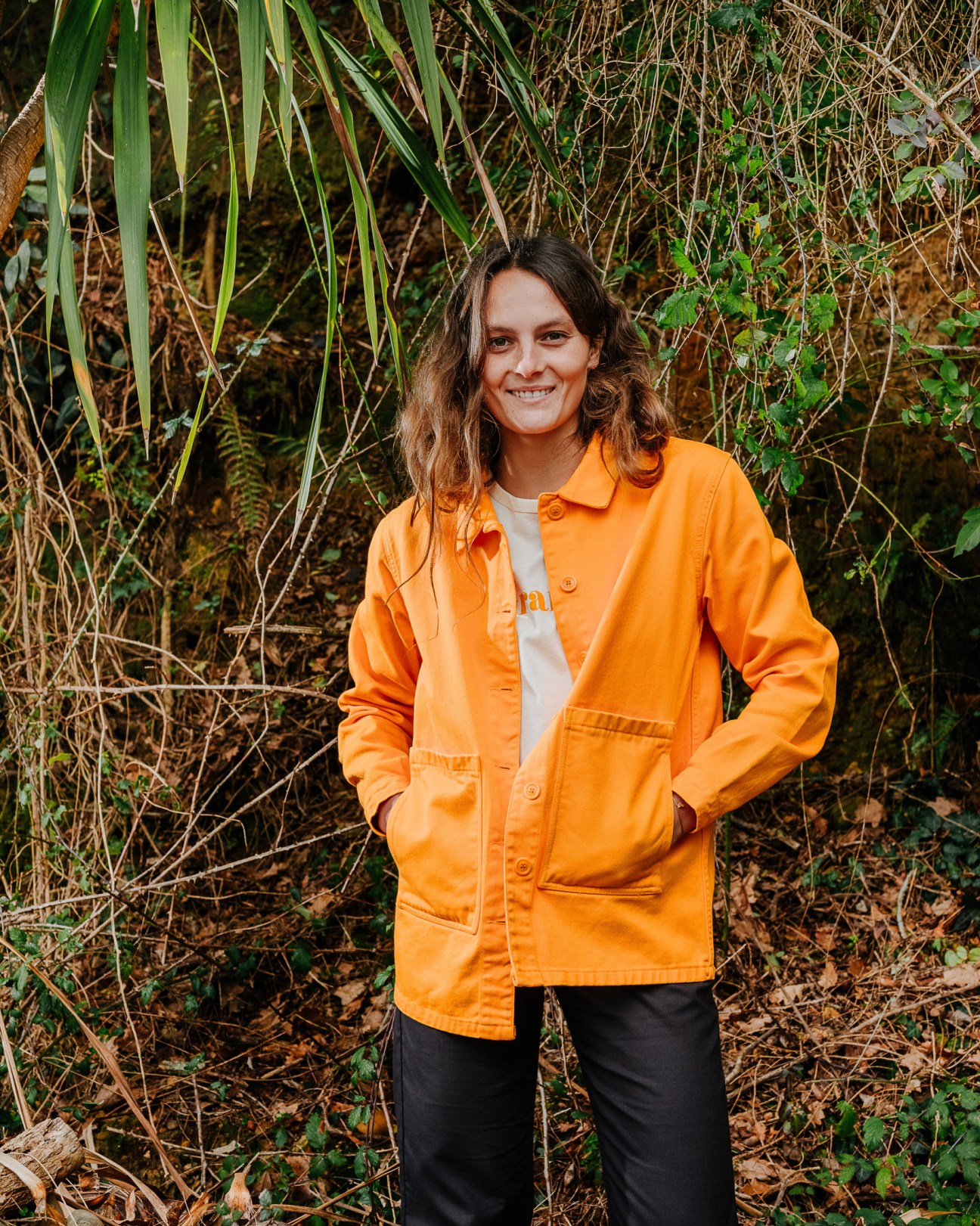 Chloé Chabaud wearing a bright-yellow French worker's jacket from her brand Graine, standing against overgrown vegetation.