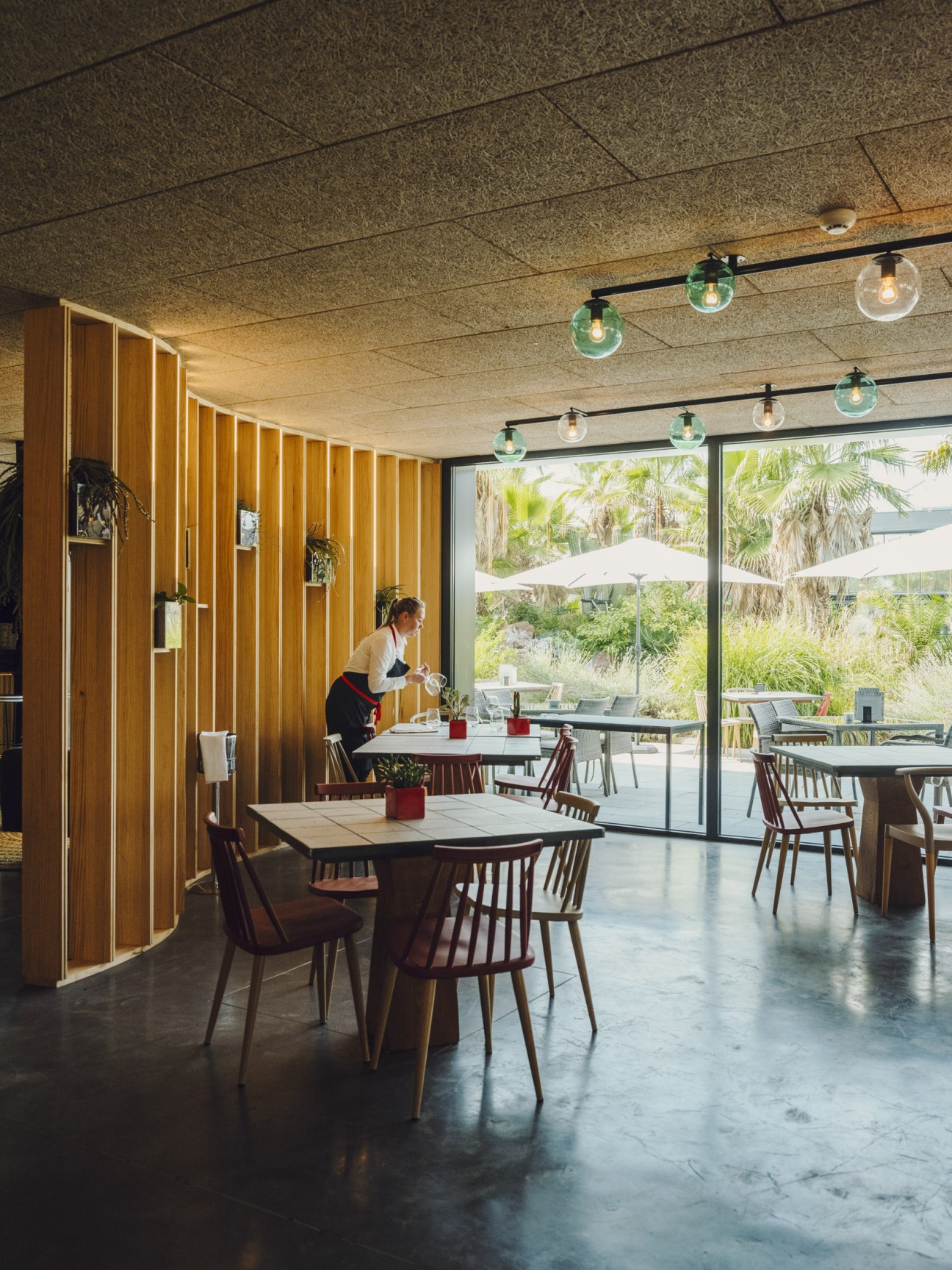 Interior of Oasis Café restaurant with wooden slat walls, pendant lighting, and views of the Desert City gardens through large windows.