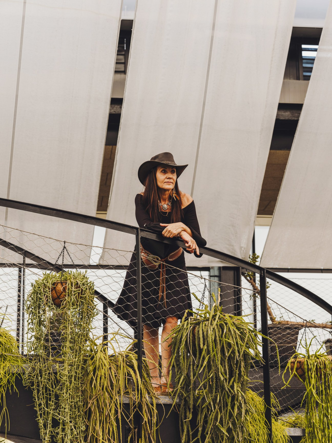 Mercedes García stands on a modern railed platform wearing a black outfit, leather cowboy hat, and beaded necklace, with hanging succulent plants visible below her and white architectural structures behind her.