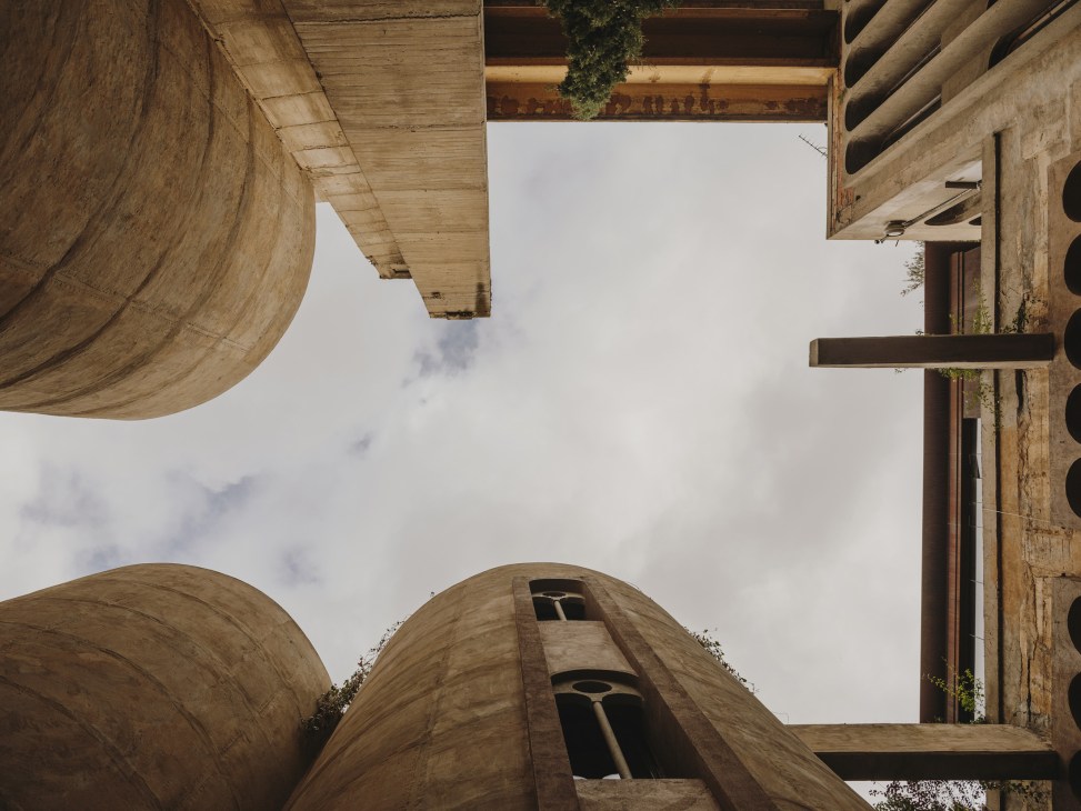 Upward view of converted concrete silos with weathered surfaces, small windows, and vegetation growing on exterior walls.