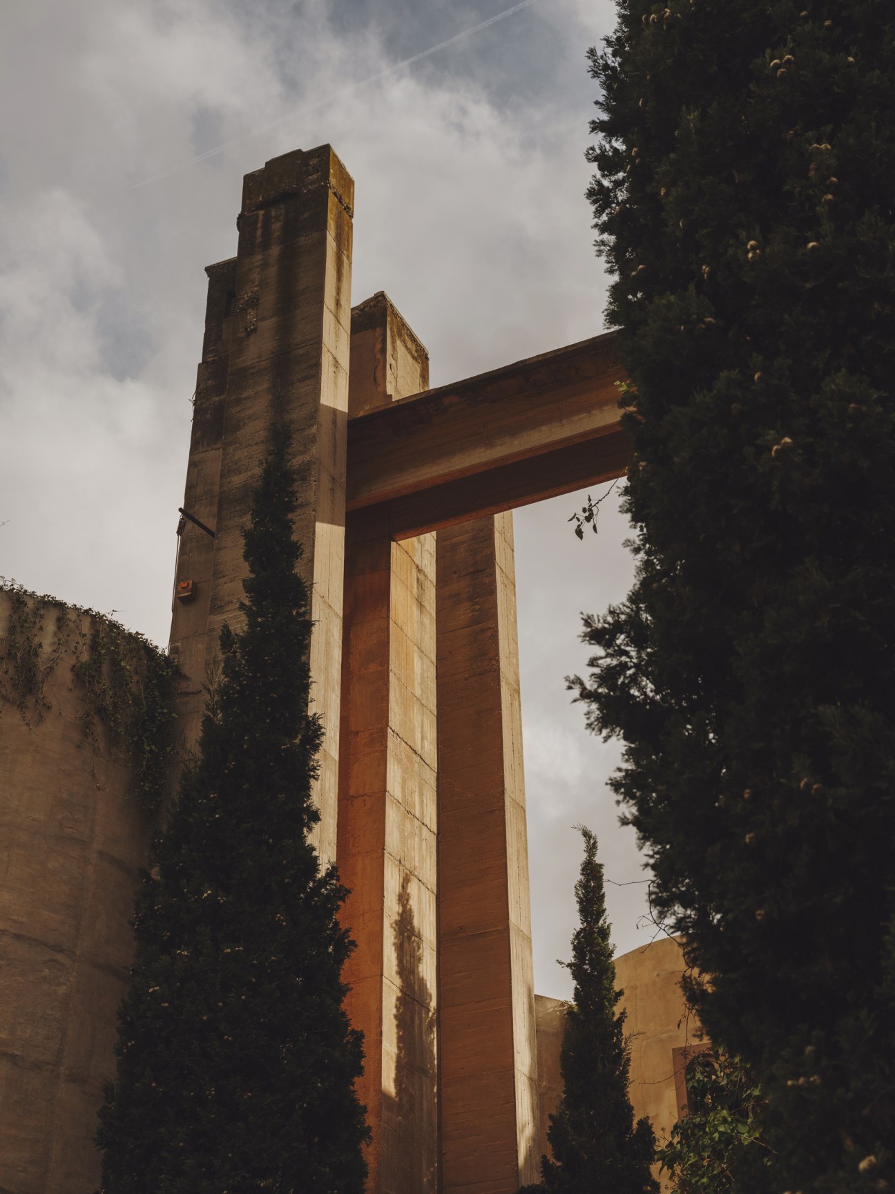 Large concrete industrial structure with ivy-covered walls and modernist architectural elements against cloudy sky.