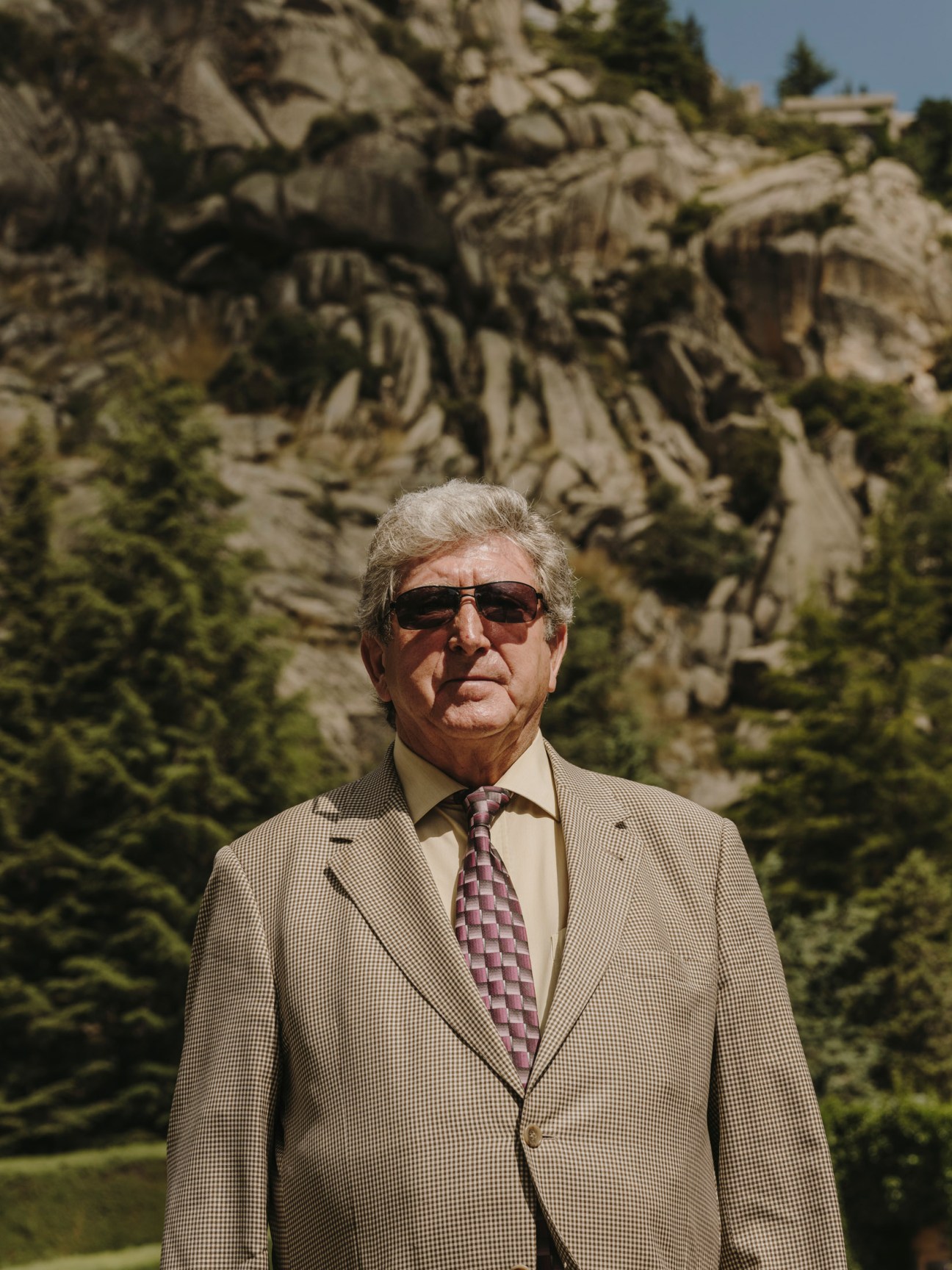 Elderly man in beige suit and sunglasses standing before rocky mountain backdrop at Valle de los Caidos