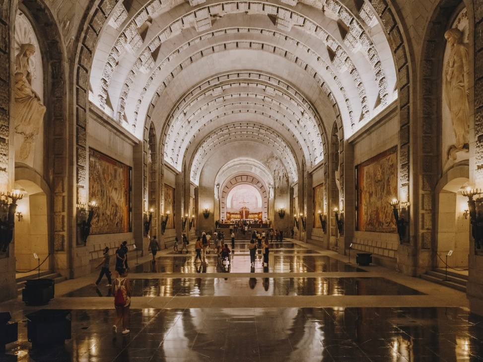 Interior of the Valle de los Caidos basilica with arched ceilings, visitors walking through the stone corridor, and altar visible in the distance.