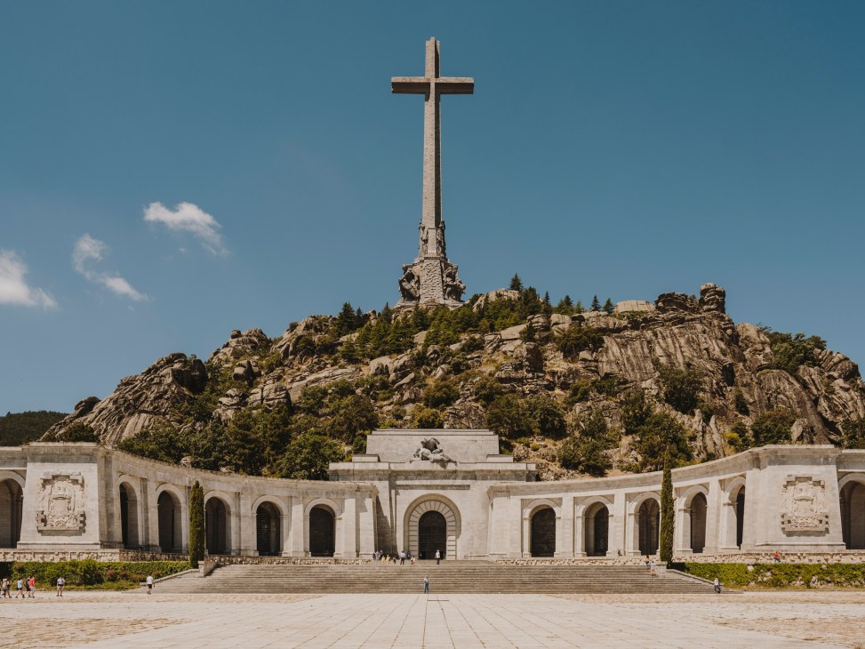 A 130-metre-tall stone cross atop a rocky mountain with a white basilica building below at Valle de los Caidos.