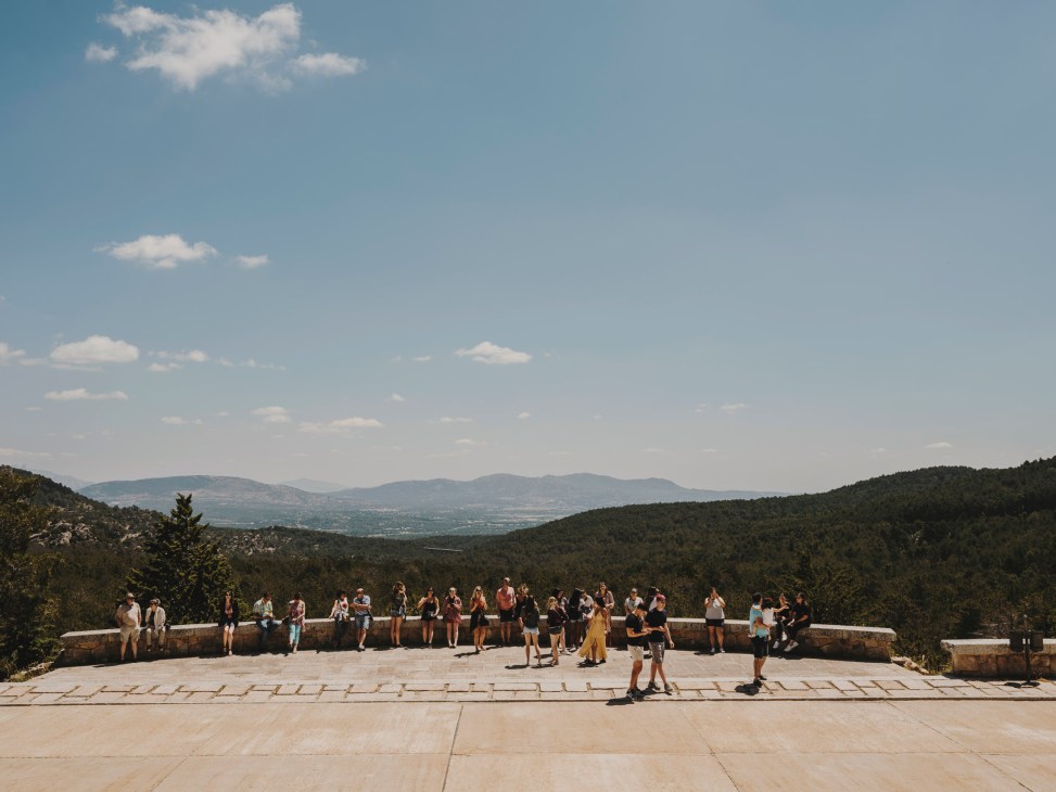 Visitors gather at a stone viewing platform overlooking a mountainous forested landscape at Valle de los Caidos monument.