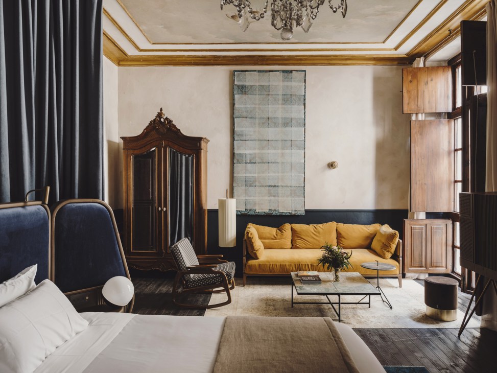 Hotel bedroom with mustard yellow sofa, navy bed, wooden wardrobe, and gold-trimmed ceiling details.