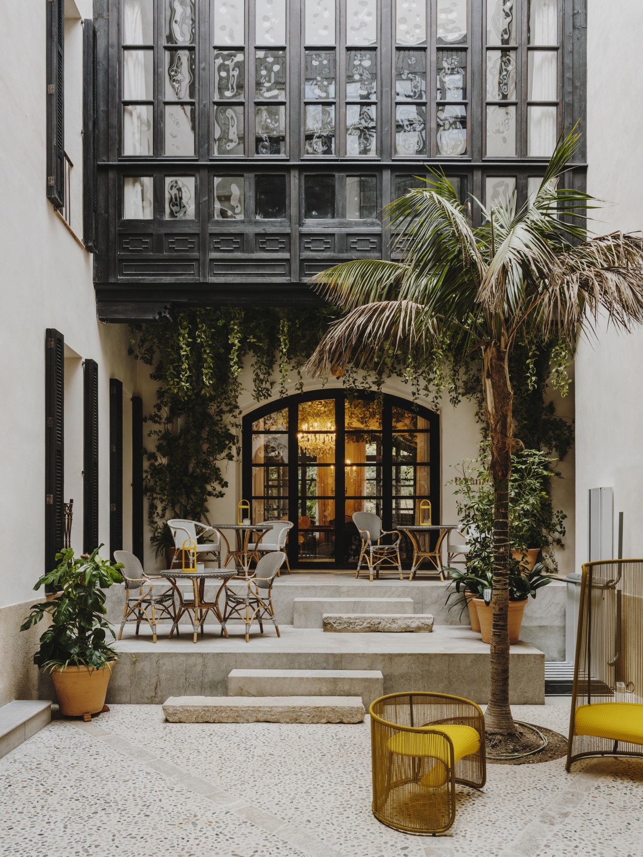 Courtyard of Can Bordoy hotel with black-framed upper level windows, palm trees, ivy-covered walls, and wicker seating areas surrounding a central terrace.