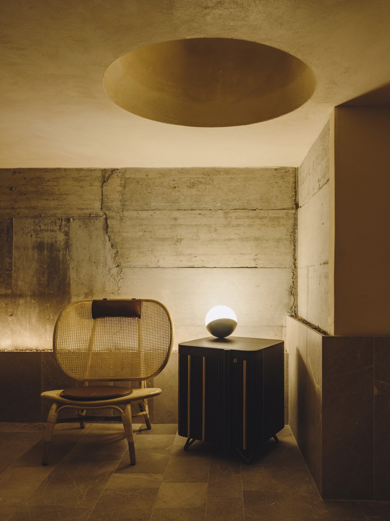 Rattan chair with black cushion and spherical lamp on dark cabinet in minimalist hotel room.