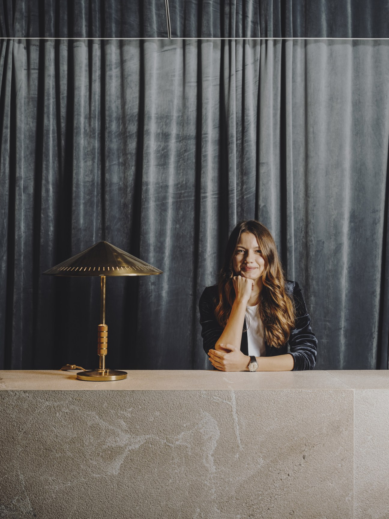 Woman in black blazer seated at desk with brass table lamp and grey curtain backdrop.