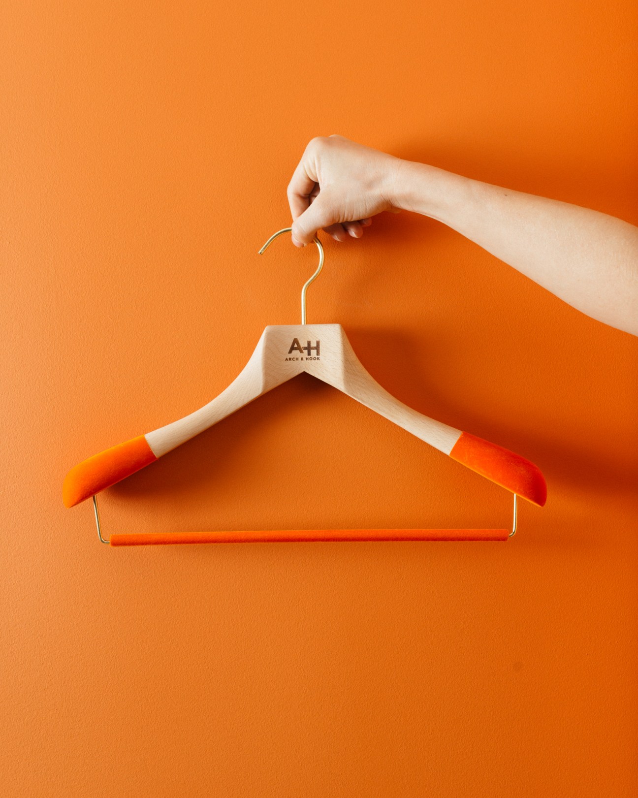Hand holding wooden hanger with orange padding labeled A+H by Arch & Hook against orange background.