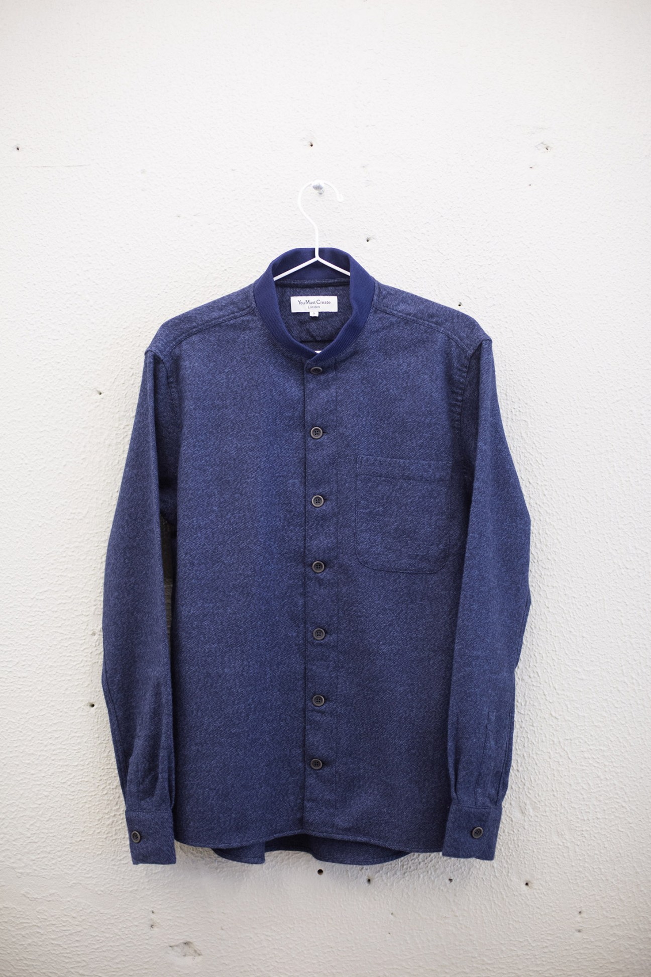 Navy blue You Must Create shirt with Mao-style collar hanging on white hanger against white wall.