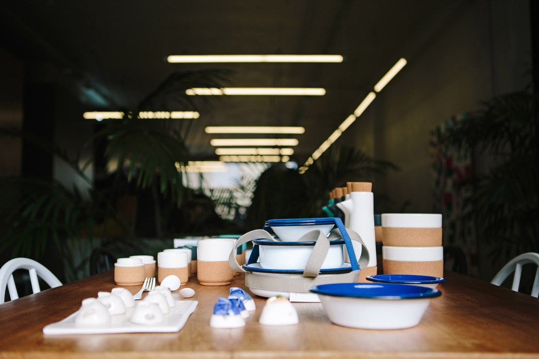 Kitchenware display on wooden table featuring white bowls with cork bands, blue handles and utensils under warm lighting.