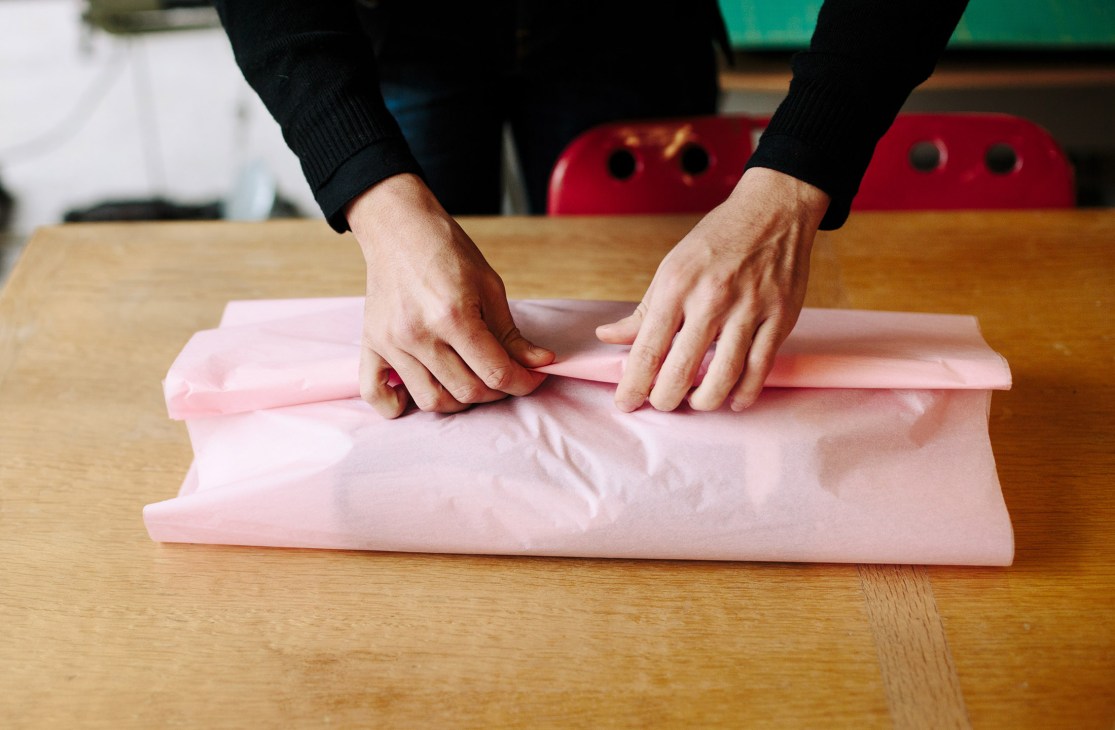 Hands folding a pink textile or garment on a wooden table in EarlyMade shop.