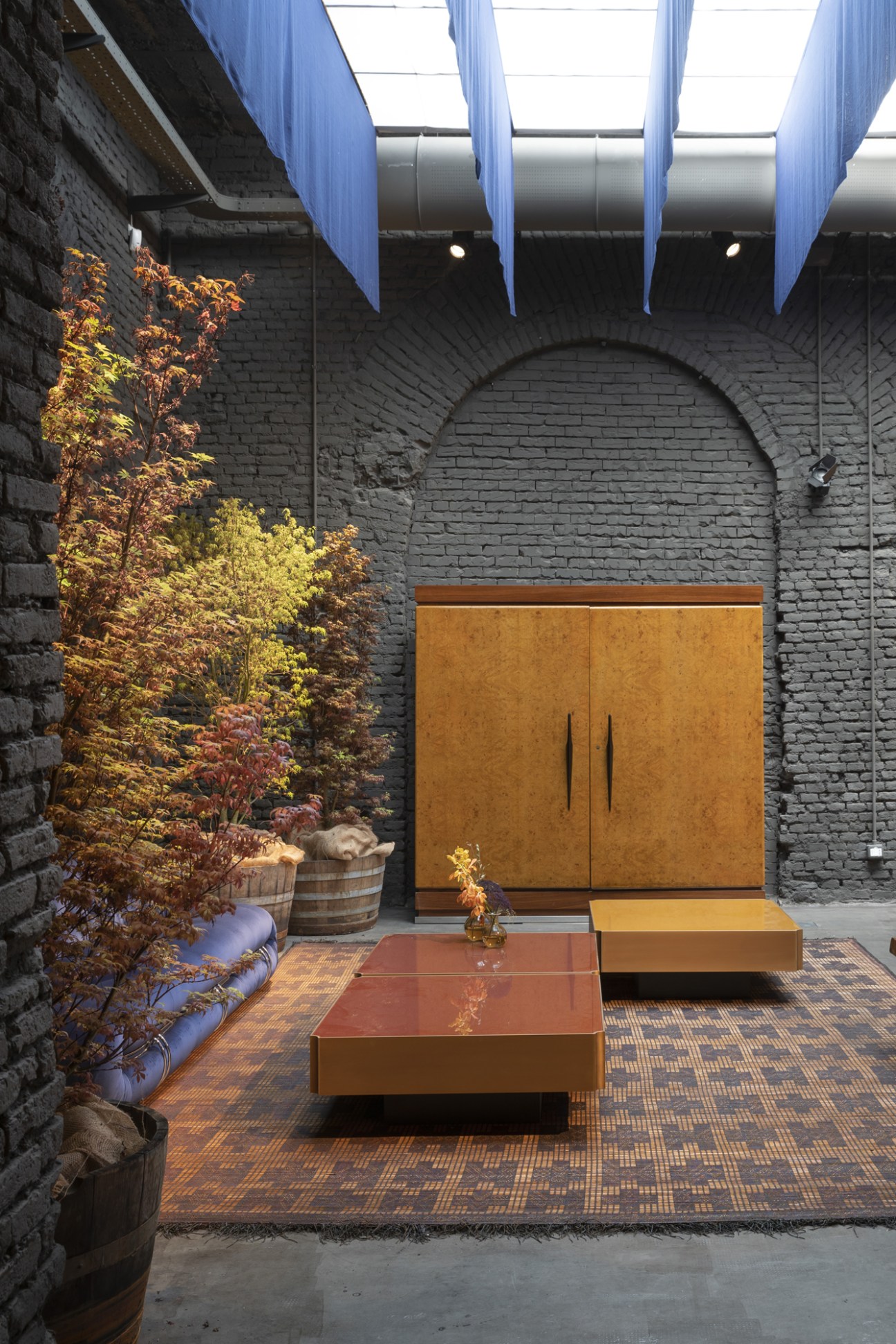 Six Gallery interior in Milan with black brick walls, autumn foliage, wooden furniture, and blue skylights overhead.