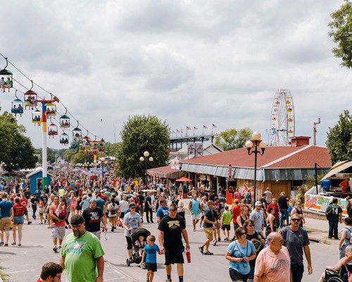 Crowds of fairgoers walking along the main avenue at Iowa State Fair with vendors, rides and attractions visible.