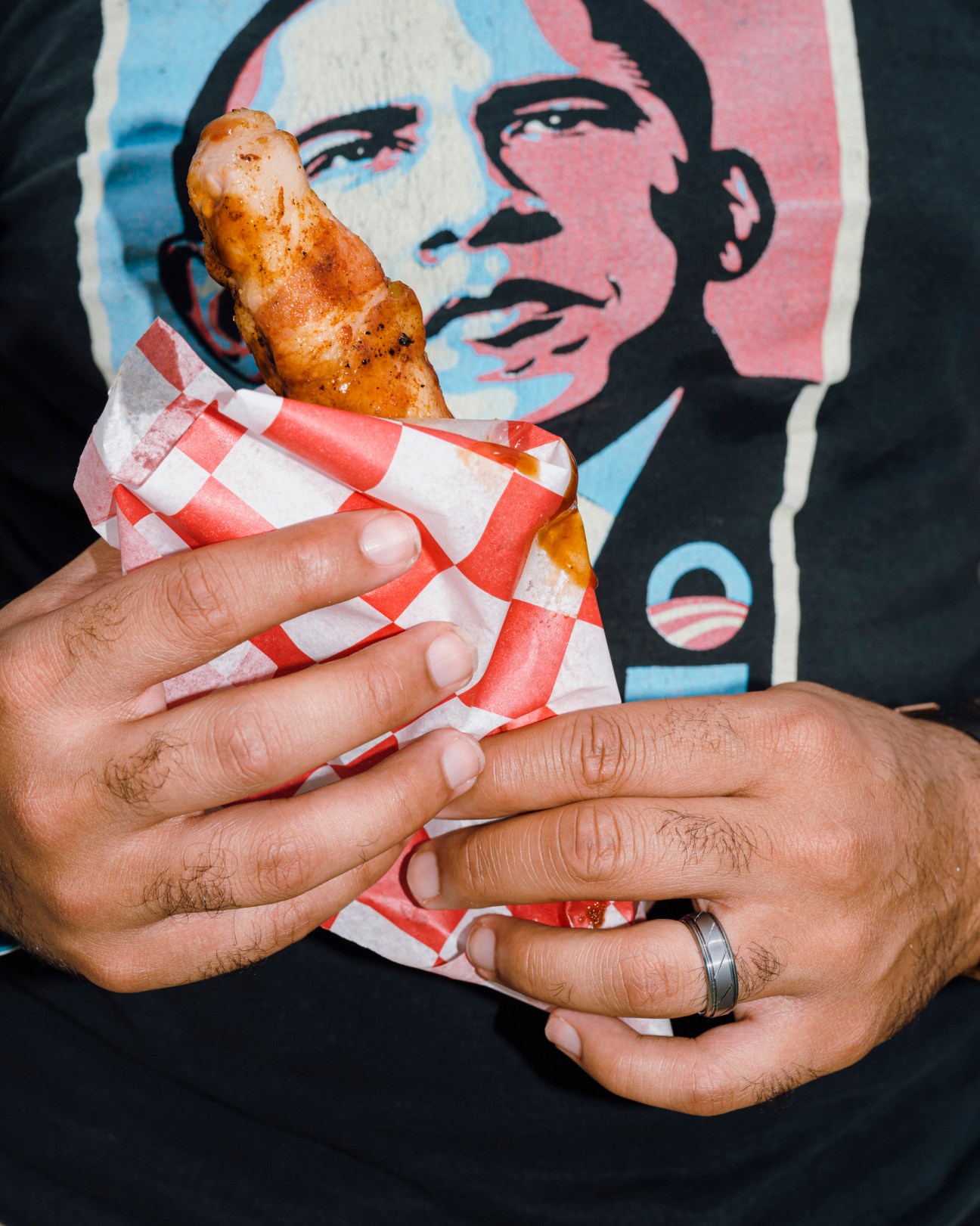 Person holding a bacon-wrapped rib in red and white checkered paper with an Obama-style portrait in the background.