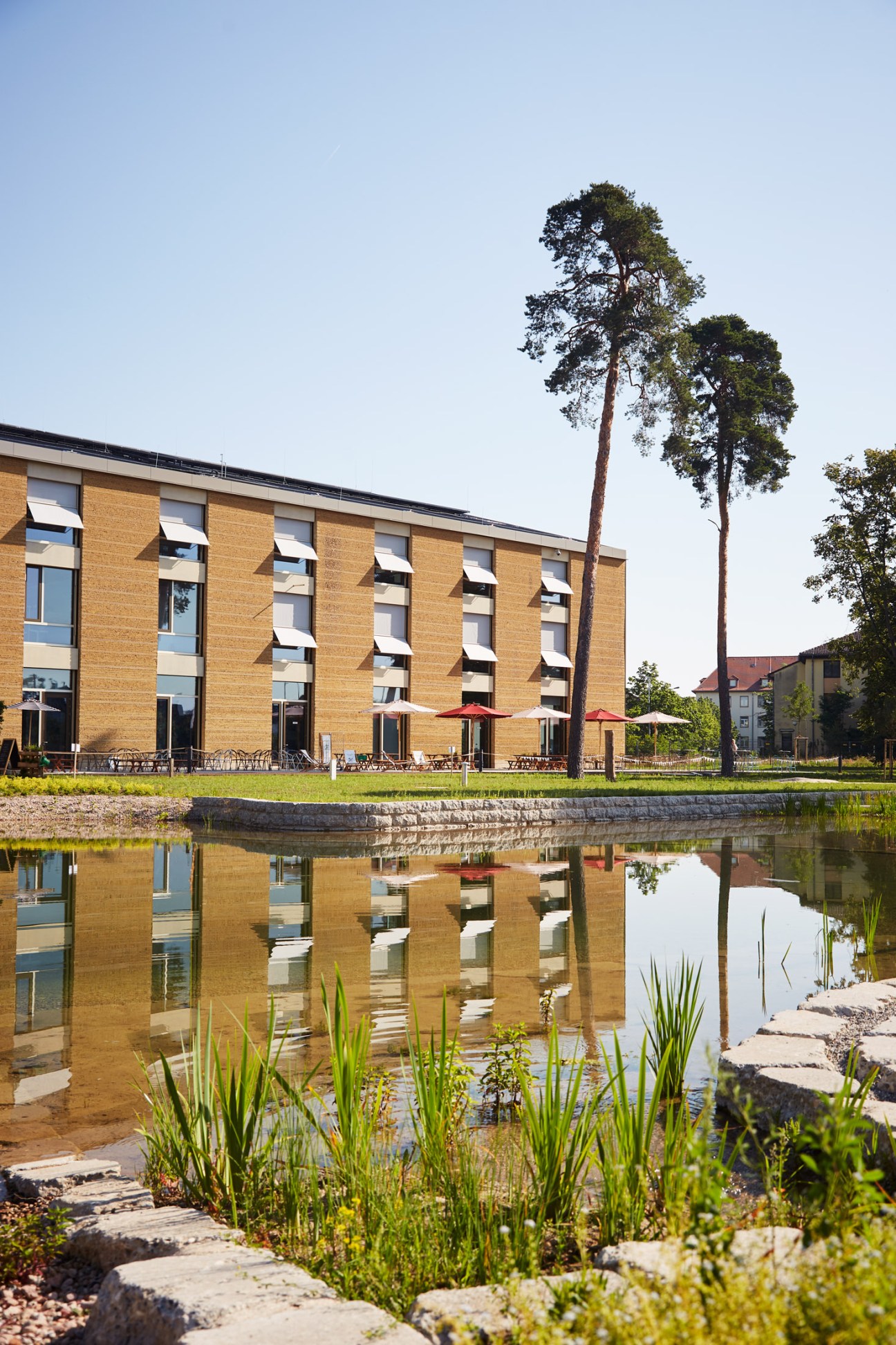 Modern brick building with large windows reflected in a pond surrounded by grass and vegetation