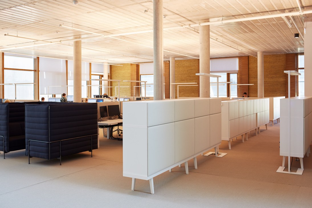 Open-plan office interior with high ceilings, natural light, wooden beams, brick walls, and modular white furniture dividers.