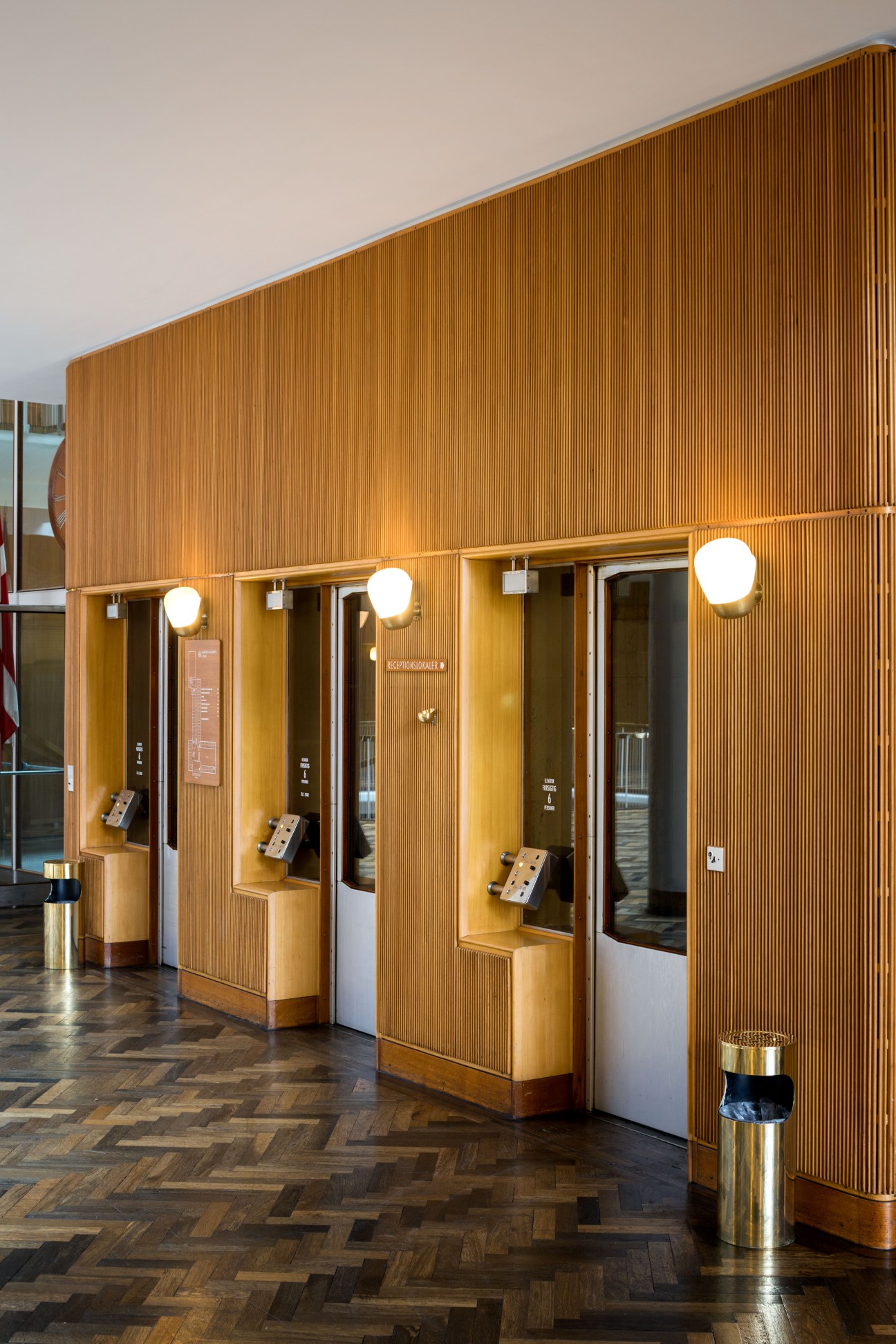 Timber-lined elevator doors with brass fixtures and round lights in Aarhus City Hall.