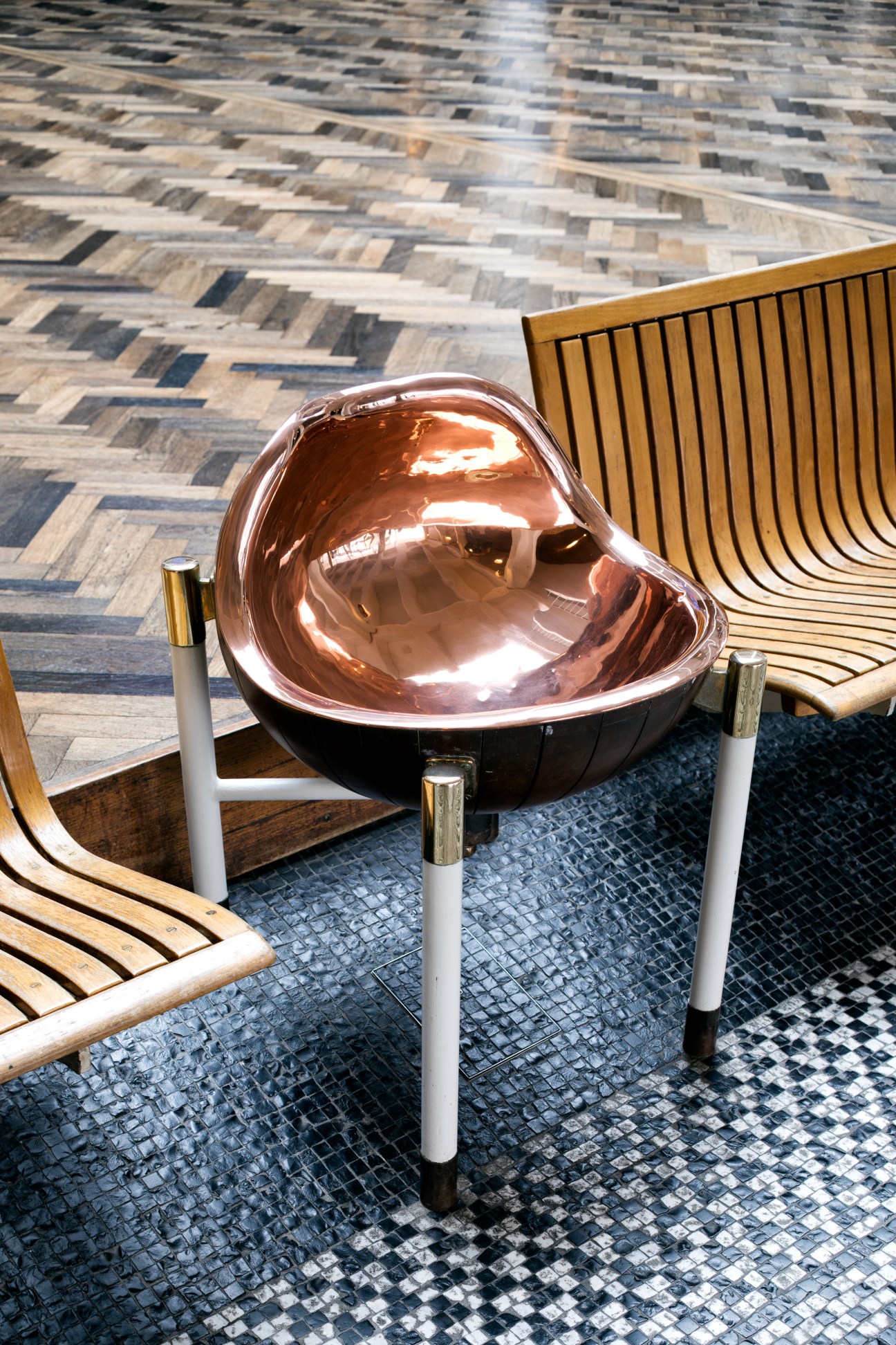 Polished brass dome fixture on white legs positioned on a geometric tile floor next to wooden chairs.