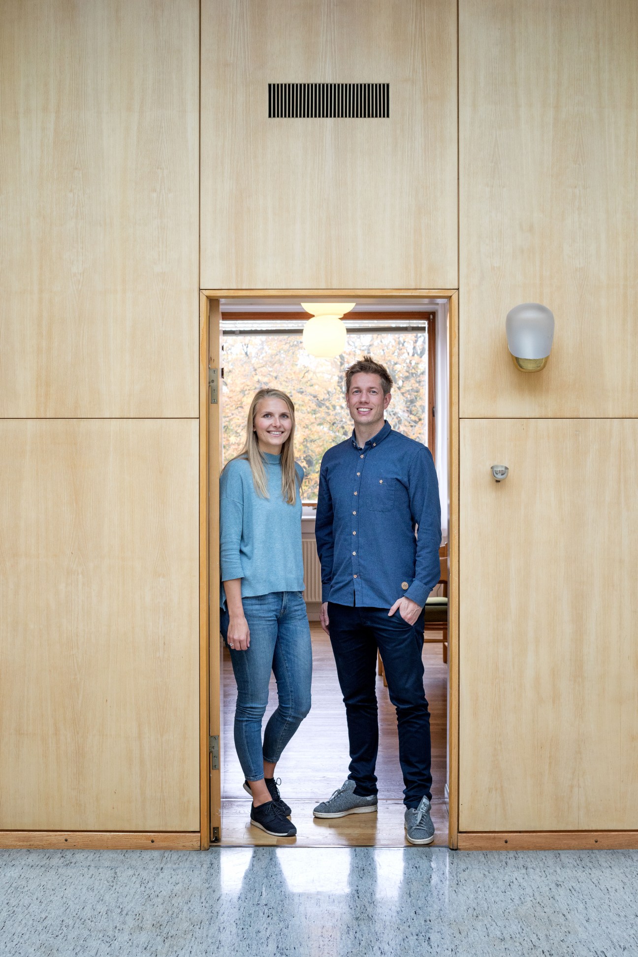 Two city hall workers stand in a timber-lined doorway with light-colored wooden walls and brass fixtures.