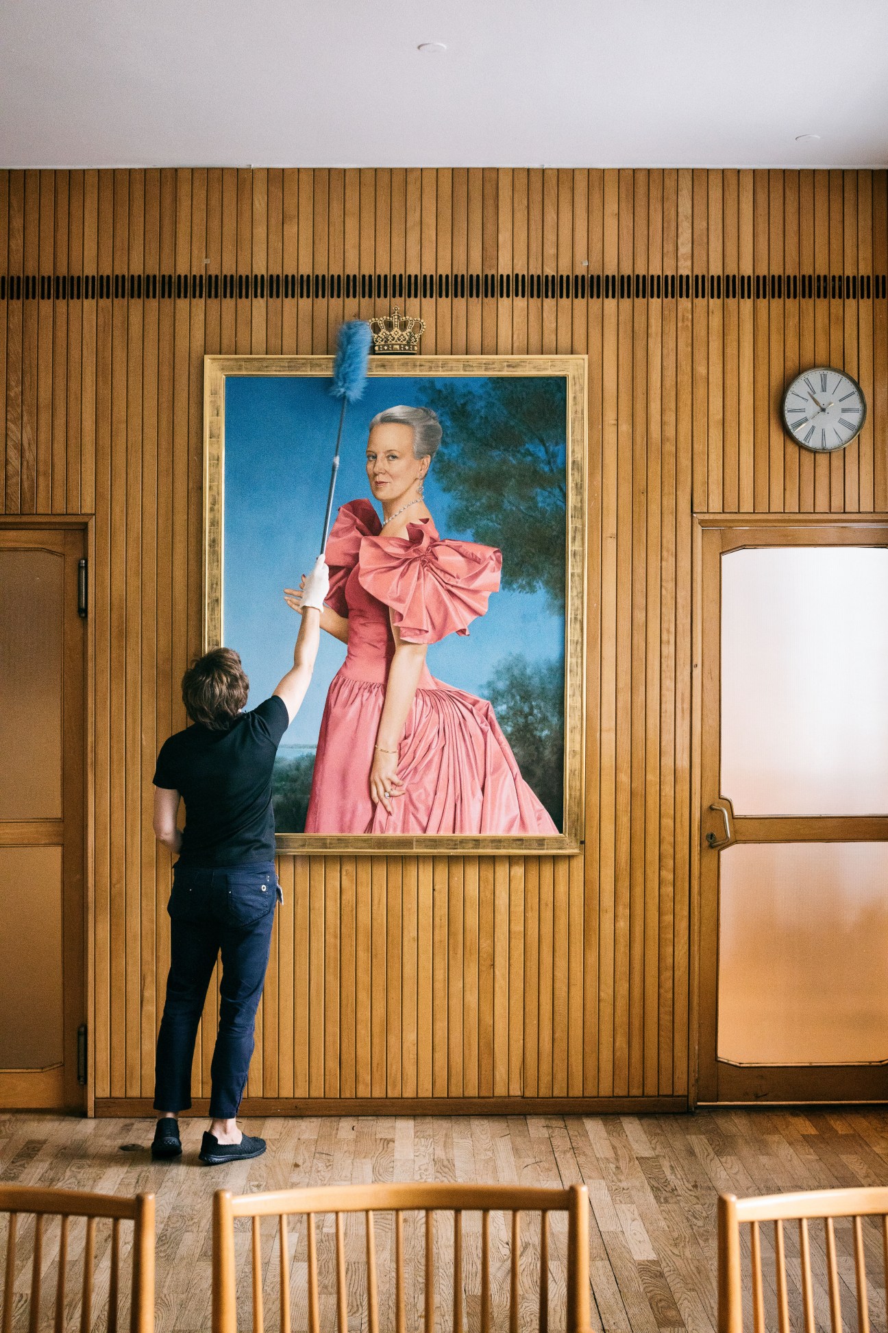 A city hall worker dusts a portrait of a woman in a pink dress hanging on a wooden-paneled wall.