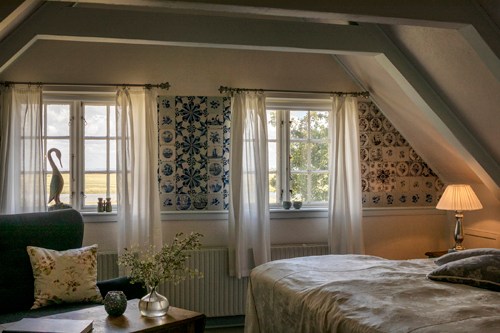 Bright hotel room with slanted ceiling, two windows with curtains, floral wallpaper, and bed with white linens