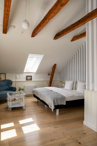Bright attic bedroom with exposed wooden beams, white walls, striped wallpaper accent, and skylight