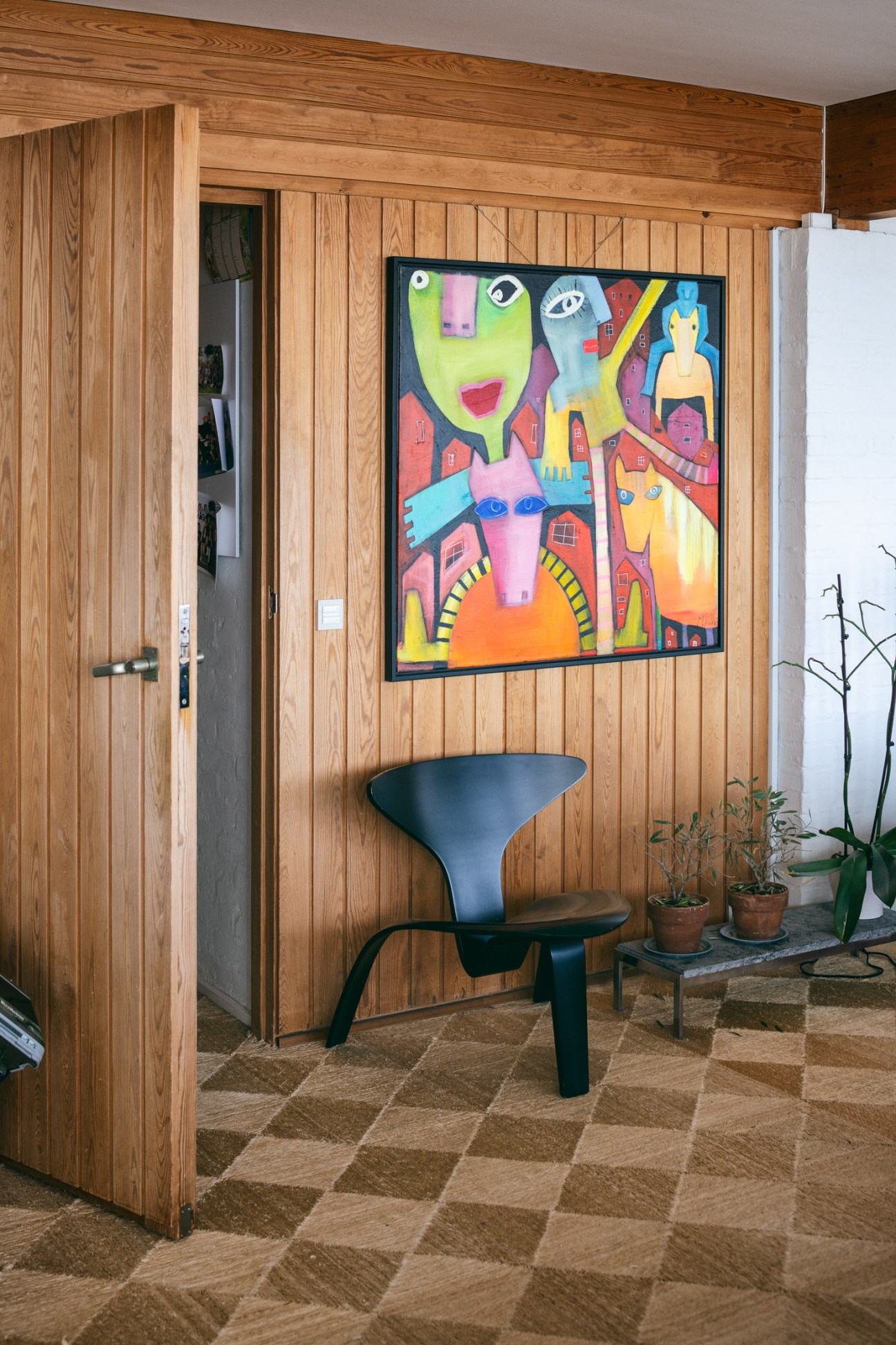 Black Poul Kjaerholm PK0 chair against natural wooden walls with colorful artwork hung above and woven seagrass flooring.