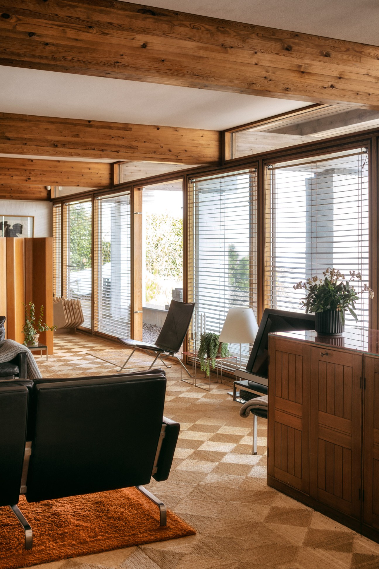 Modern living room with wooden beams, black leather Kjaerholm furniture, and panoramic windows overlooking water.