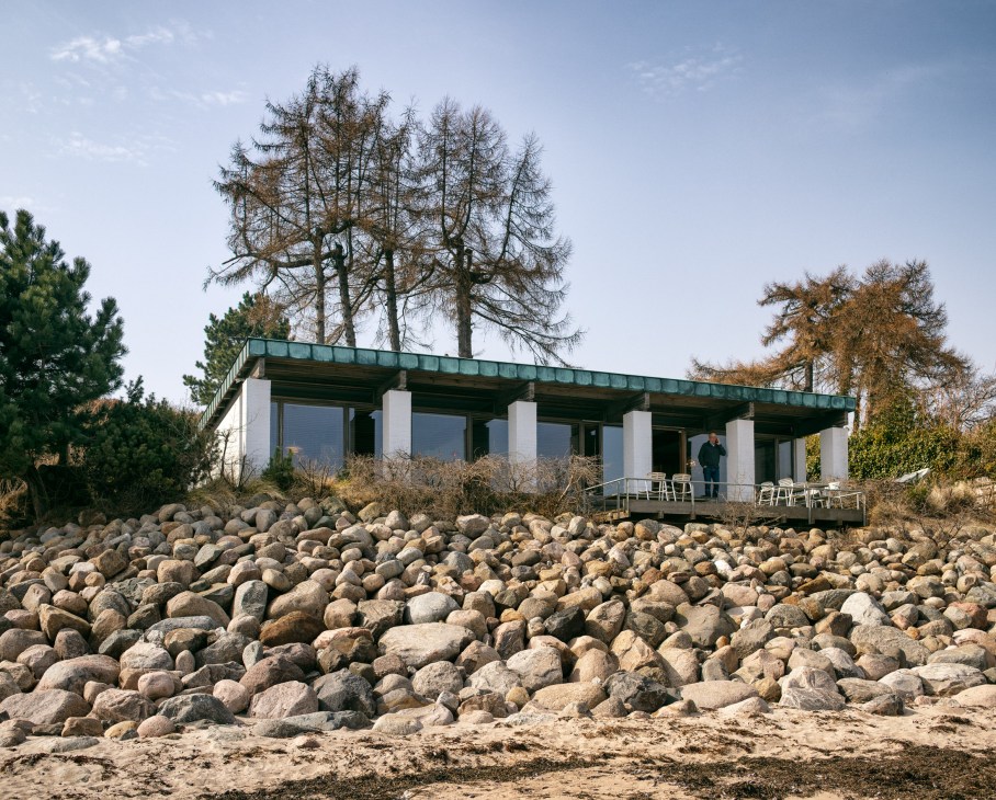 Modernist beach house with white columns and green roof overlooking rocky shore and Øresund strait.