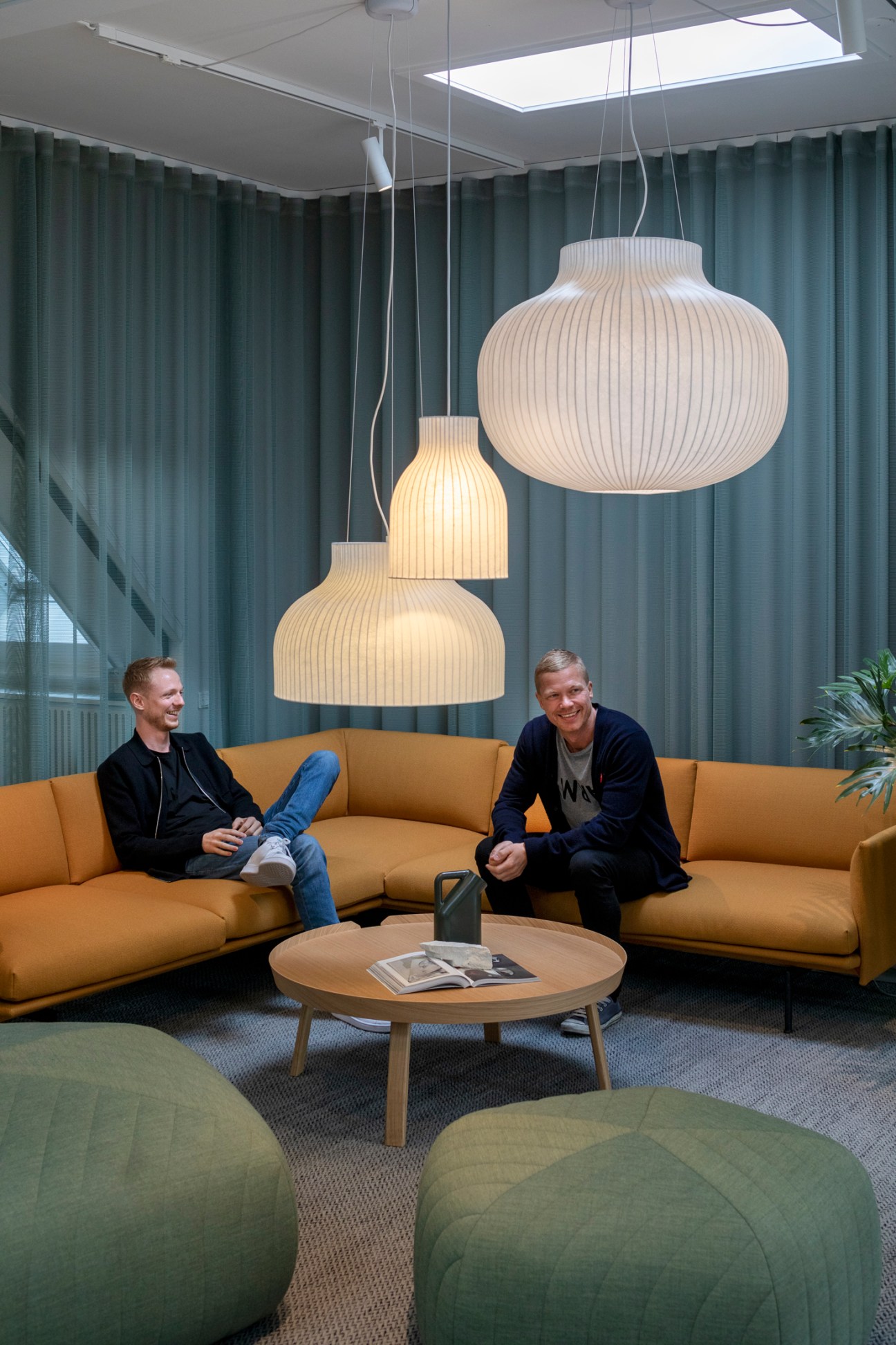Two men sitting on a mustard yellow sectional sofa beneath pendant lights in a modern office space